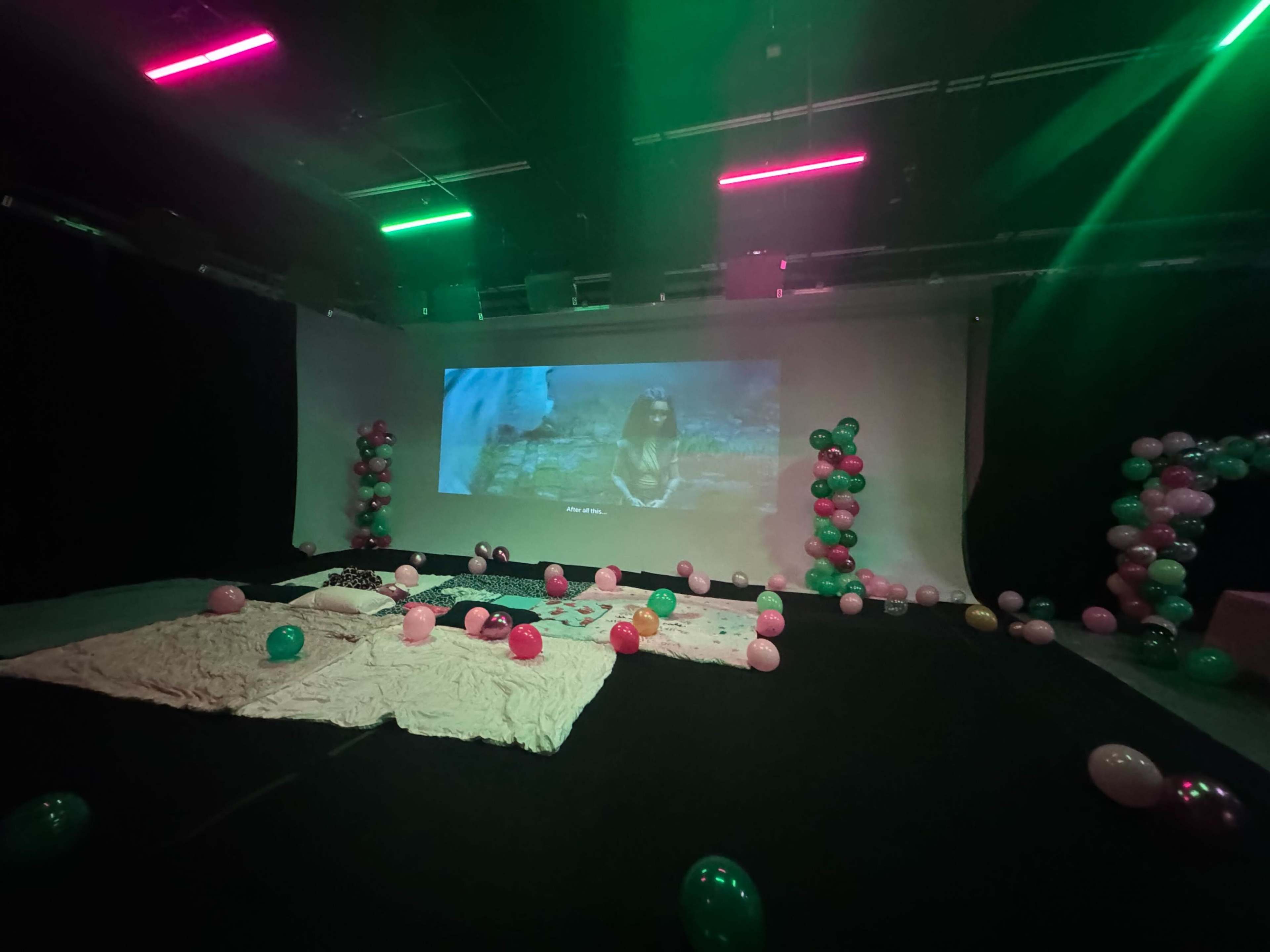 The scene shows a dimly lit room with a large screen displaying a film, surrounded by pink and green balloons and blankets scattered on the floor.