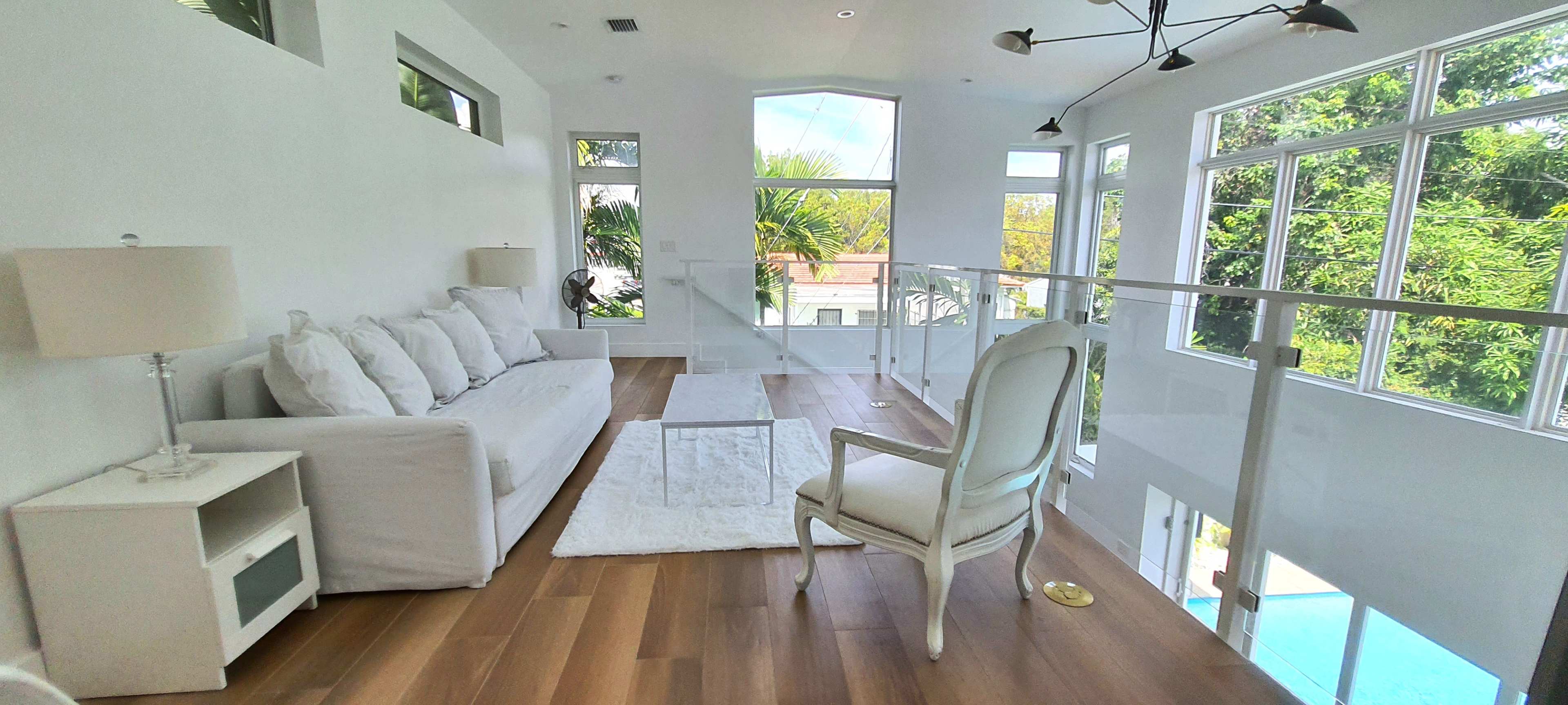 A bright, modern living area features a white sofa and a glass coffee table, with large windows overlooking greenery and a pool.