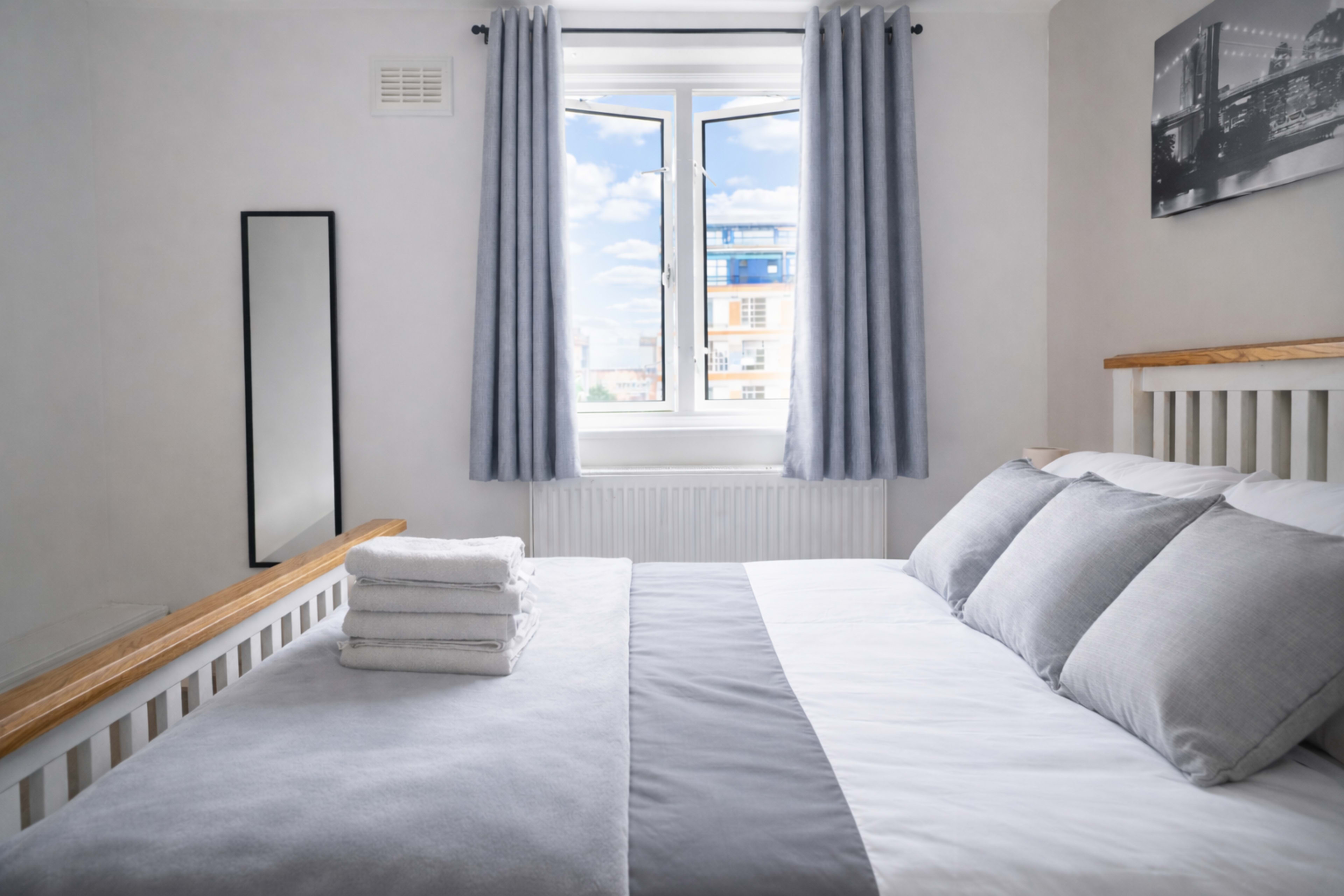 A neatly made bed with folded towels sits in a well-lit bedroom featuring a large window and light-colored curtains.