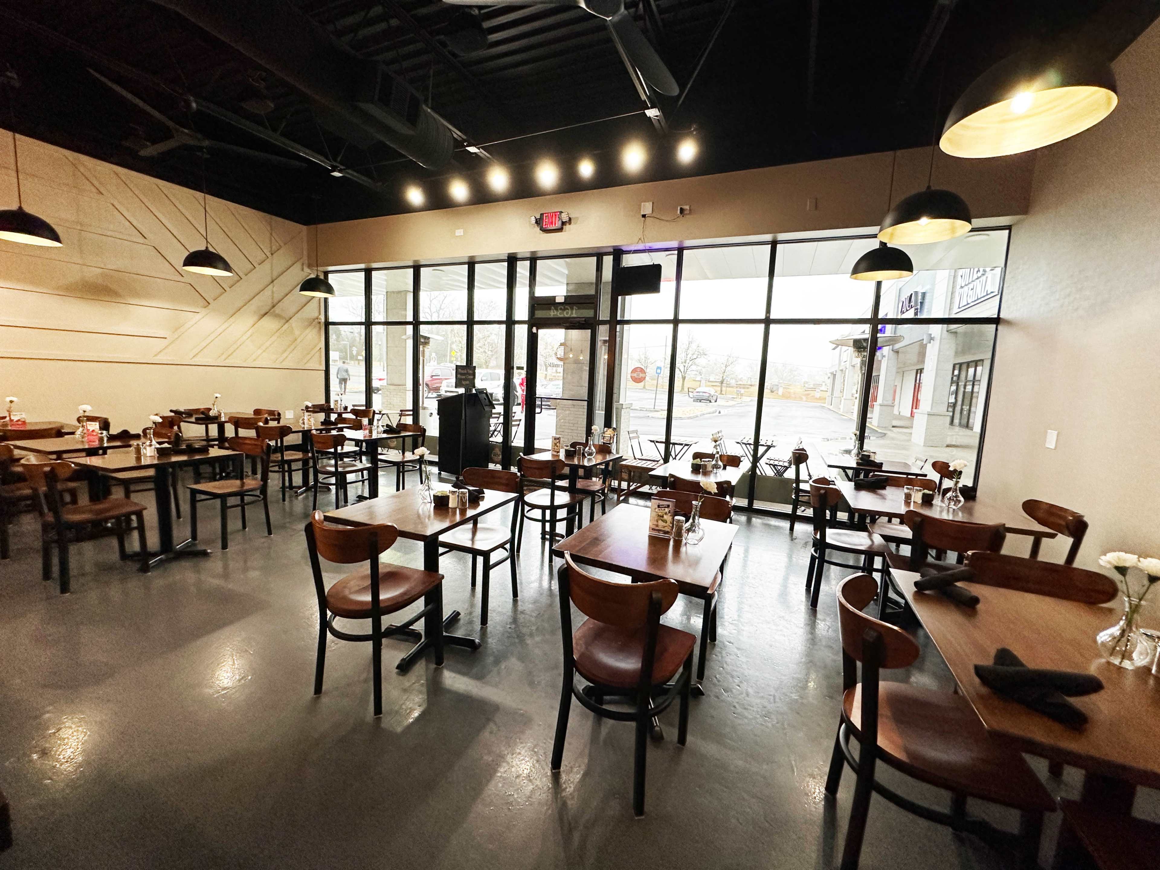 A modern restaurant interior with several wooden tables and chairs, large windows allowing natural light, and a clean, minimalist design.