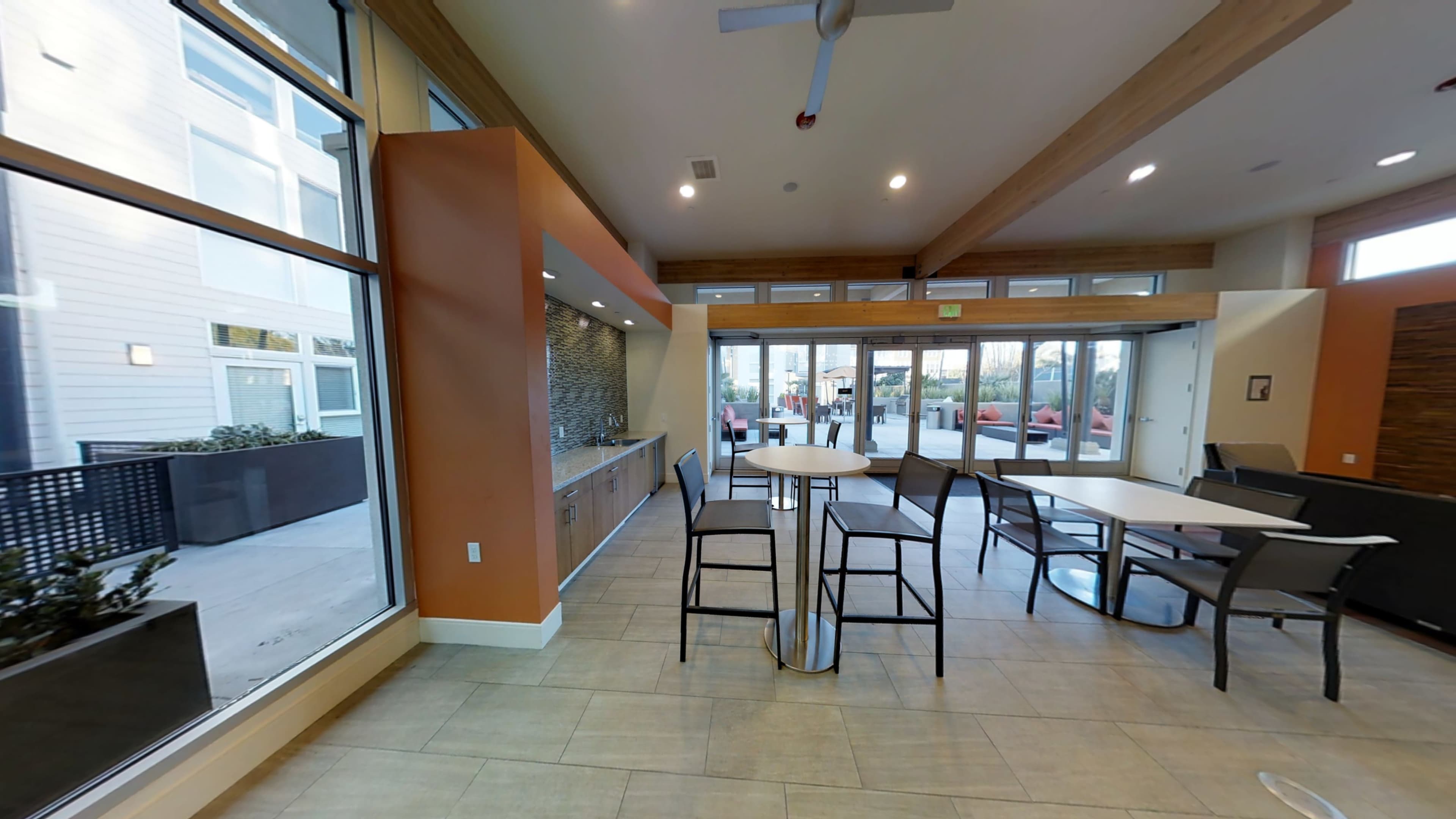 A modern indoor communal area with tall tables and chairs, large windows, and a kitchen space along one wall.