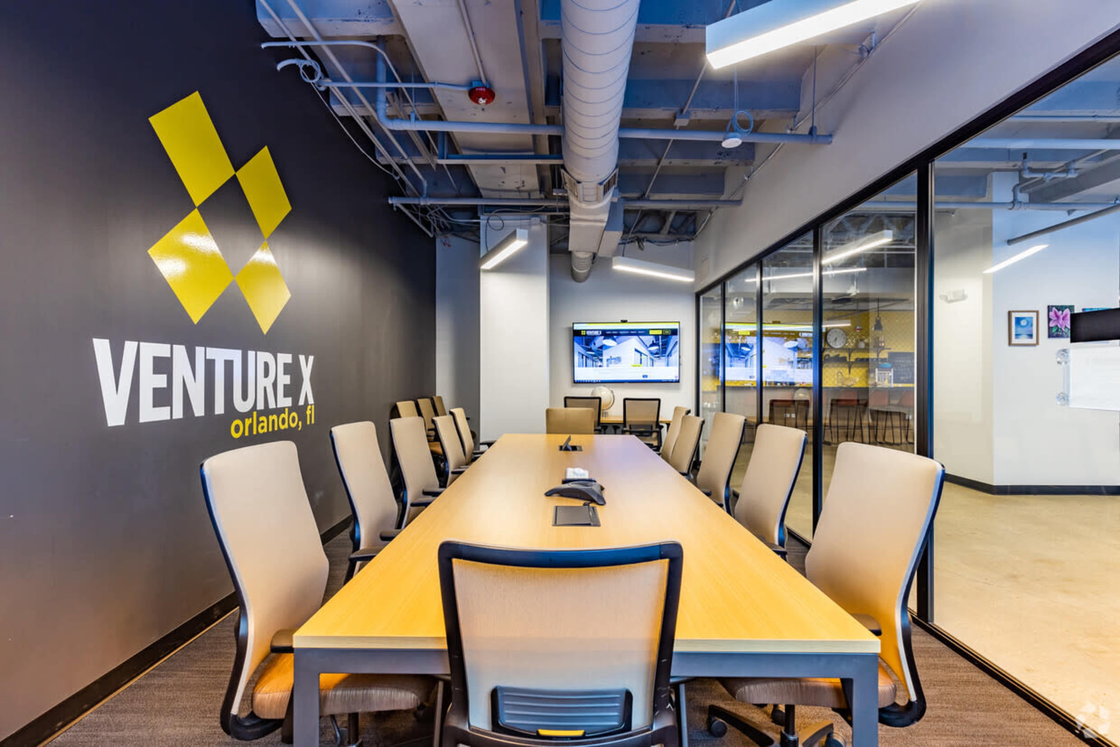 A modern conference room features a long wooden table surrounded by ergonomic chairs, with a large wall displaying the "Venture X" logo.
