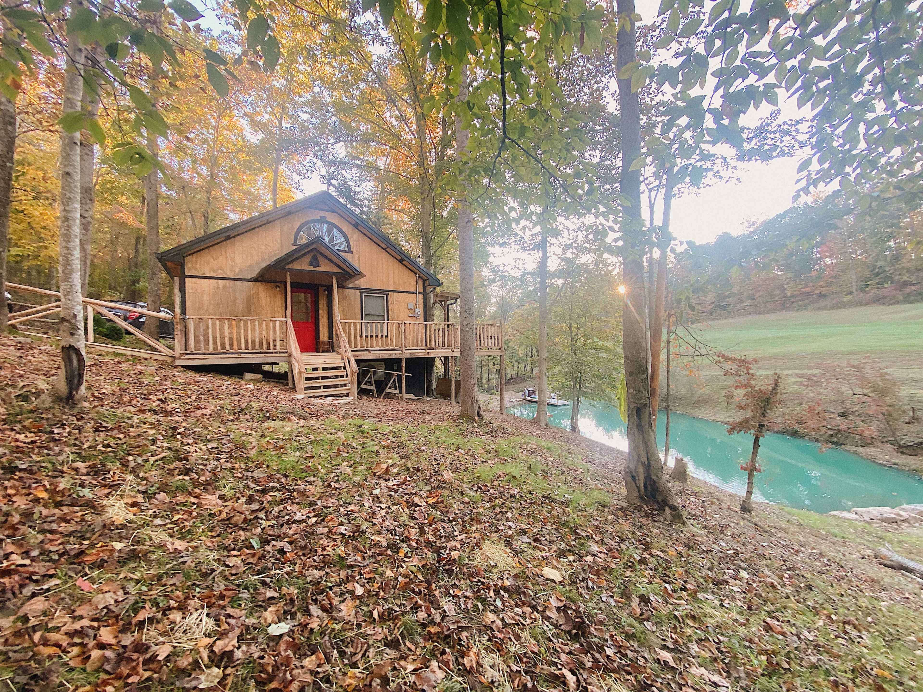 A wooden cabin with a red door is situated by a turquoise pond, surrounded by trees with autumn foliage.
