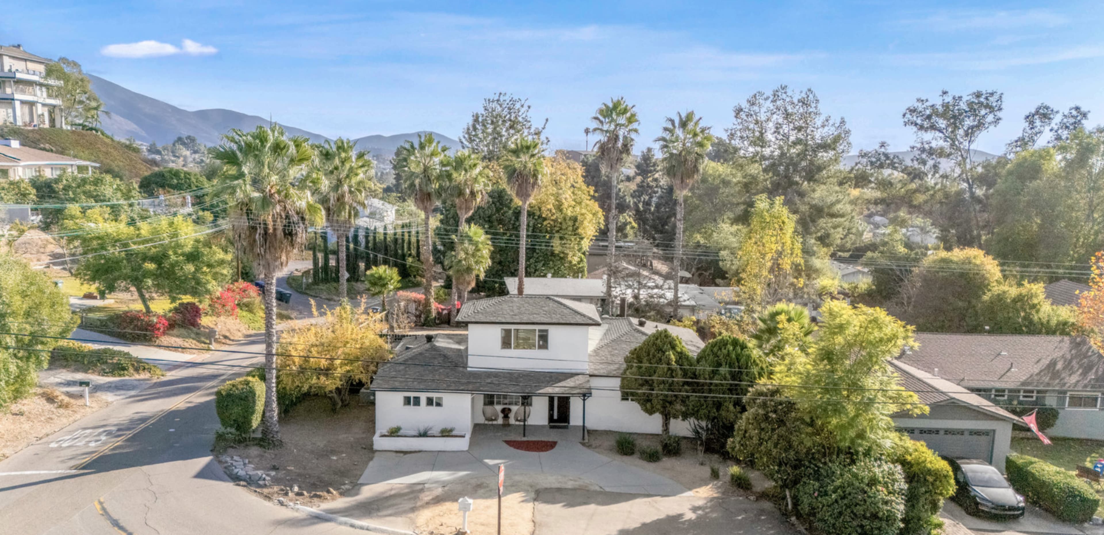 The image shows a two-story house surrounded by palm trees and lush greenery, located on a corner plot in a residential area.