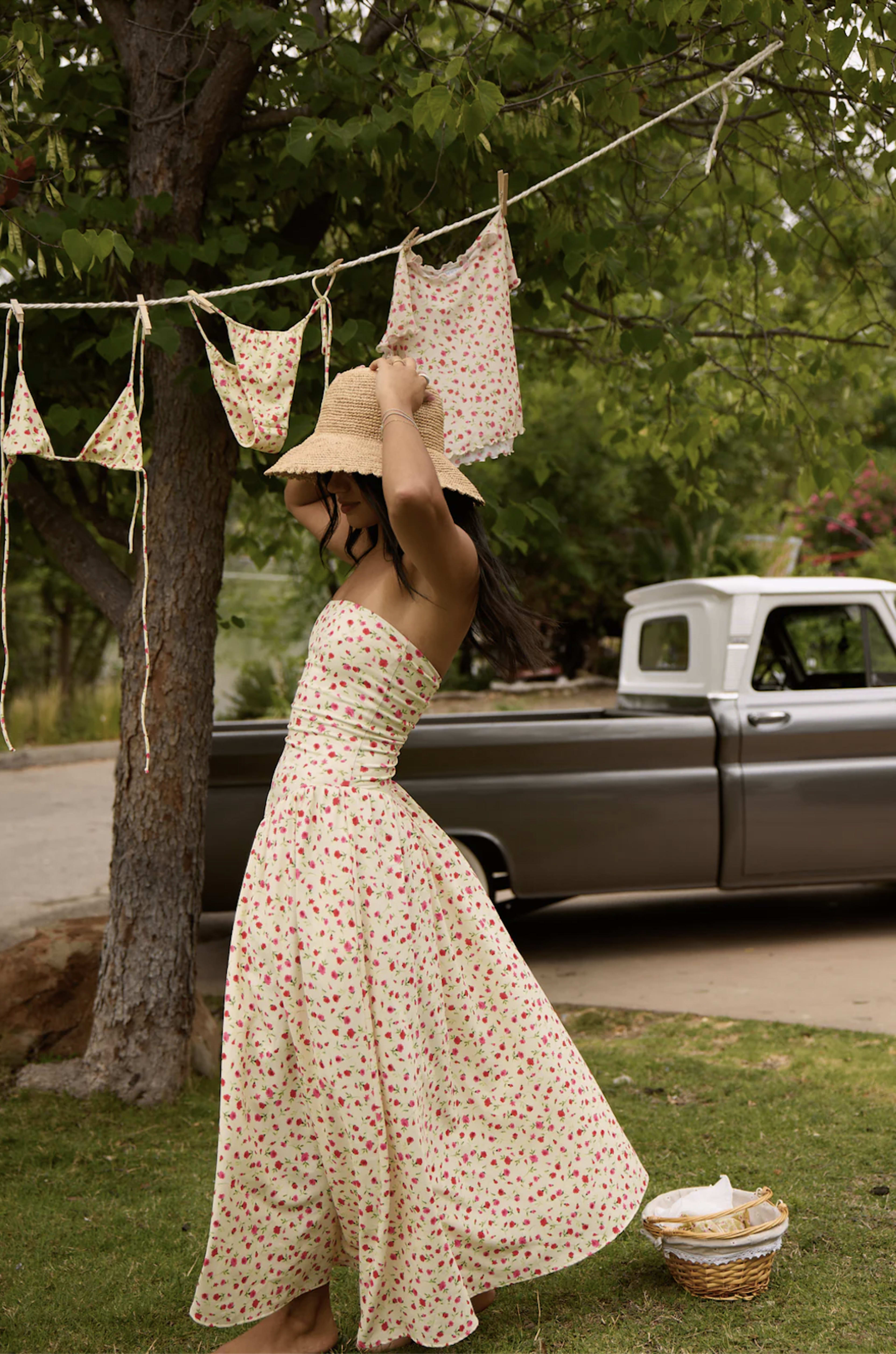 A woman in a floral strapless dress twirls under a tree near a vintage pickup truck, with laundry hanging on a clothesline.