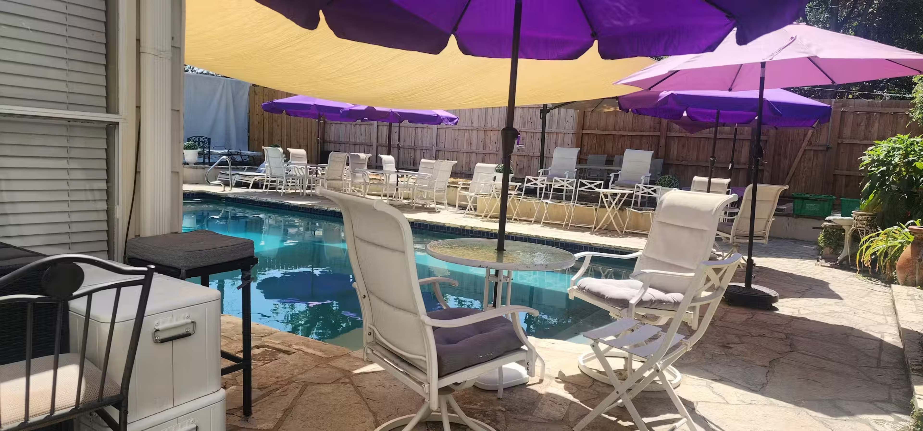 A pool area is shaded by purple umbrellas and features white lounge chairs arranged around the water.