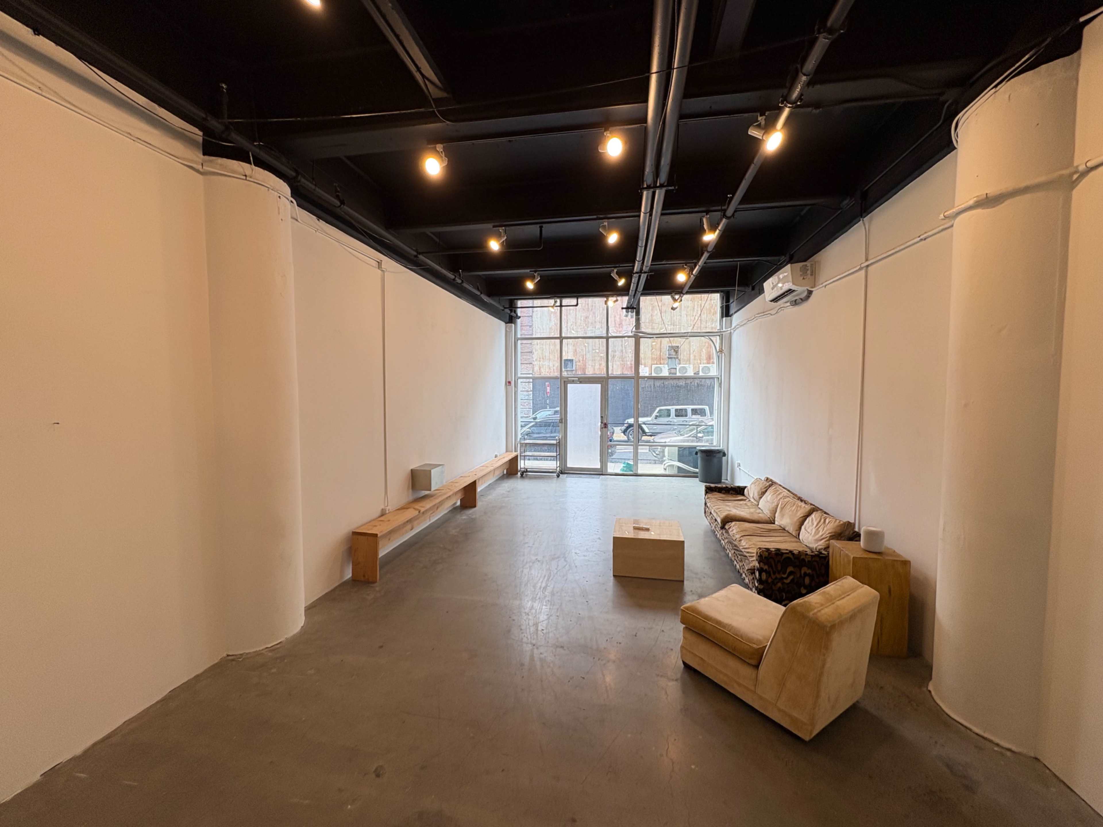 The image shows an empty interior space with white walls, concrete flooring, and a seating area consisting of a sofa and armchair, along with a wooden bench and table.