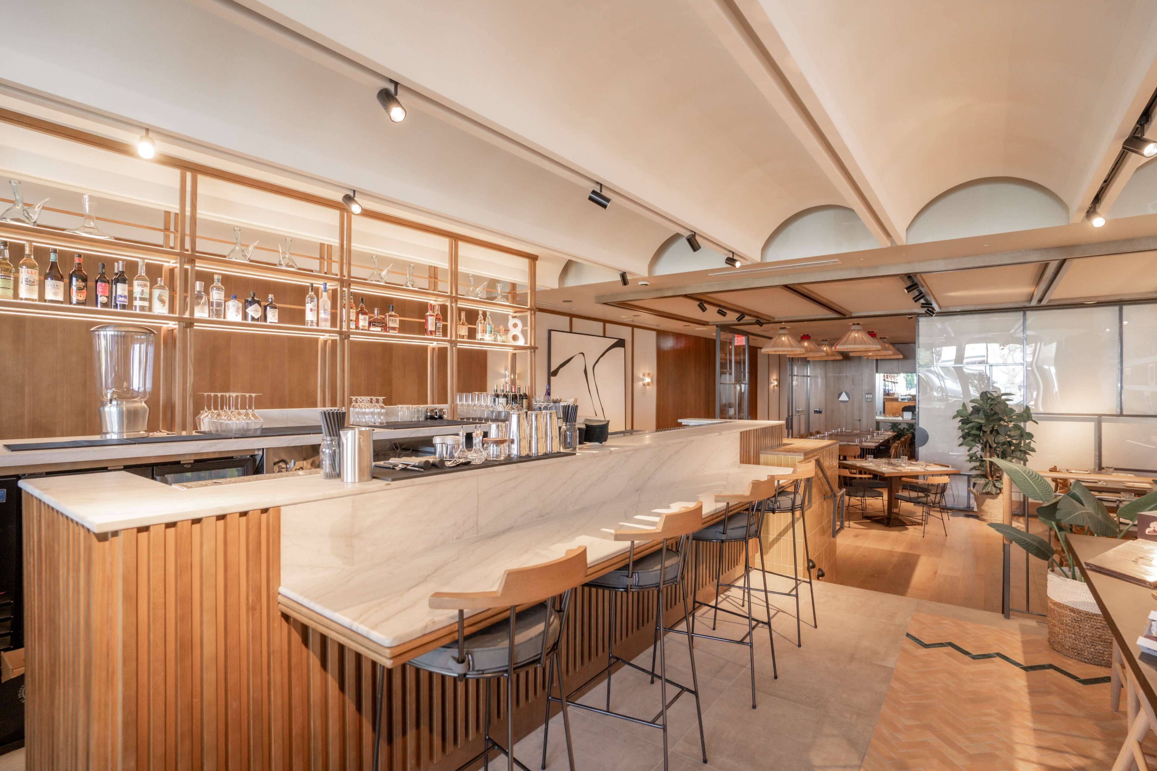 The image shows a modern restaurant interior featuring a sleek bar with a marble countertop, wooden accents, and seating along the bar.