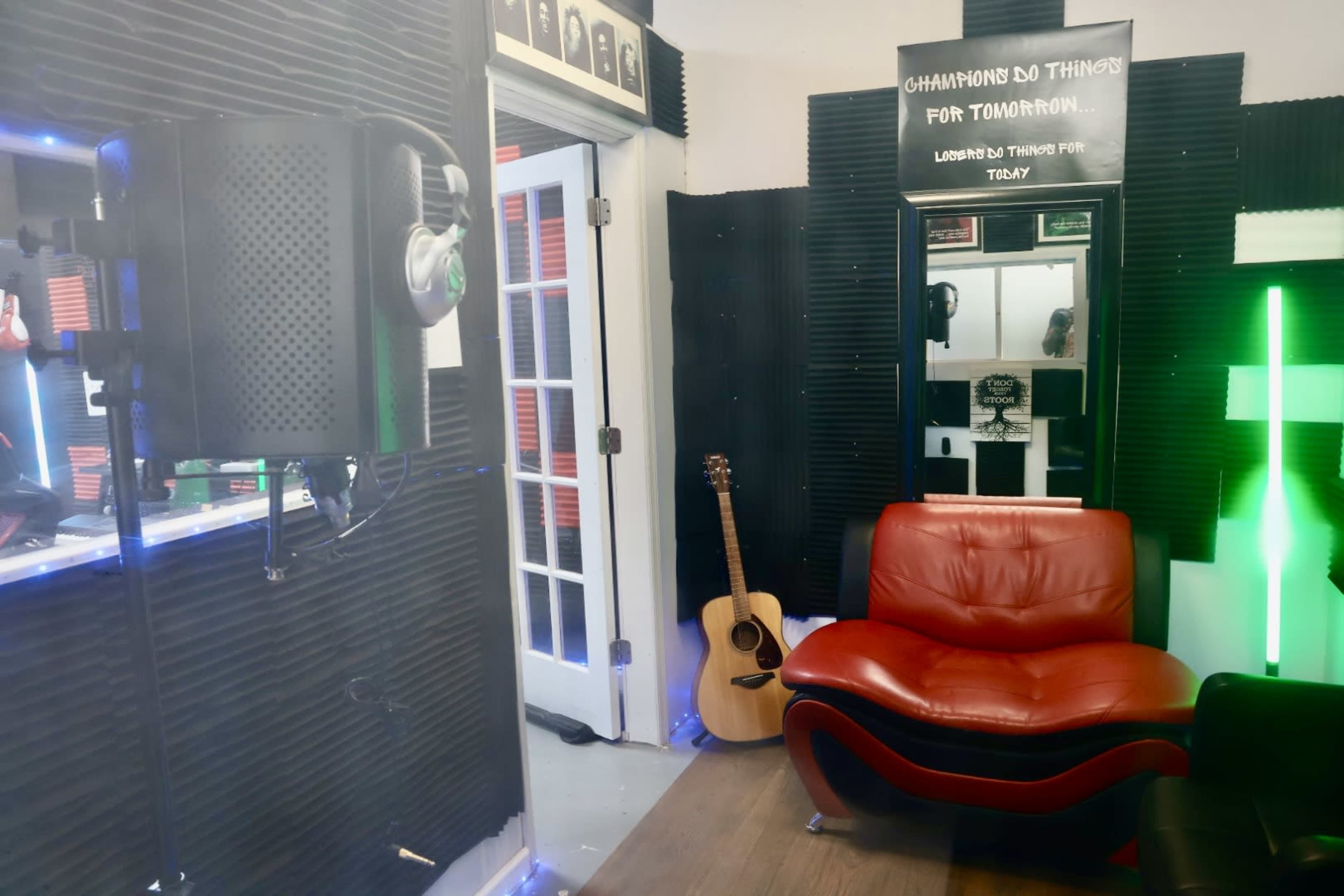 The image shows a small music studio featuring a red chair, an acoustic guitar, and soundproofing panels on the walls.