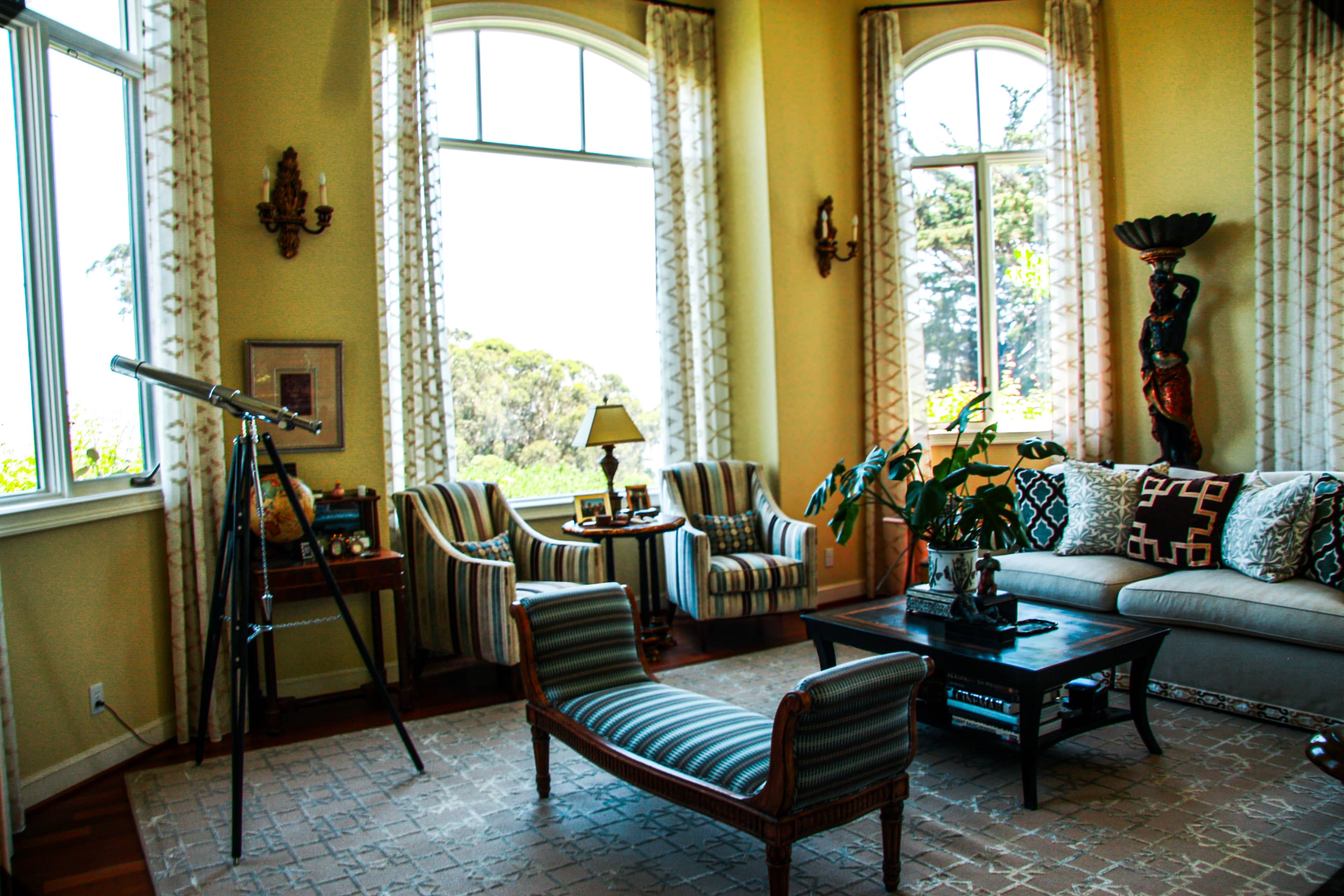 The image shows a spacious living room with large windows, featuring a telescope, two striped chairs, a couch with decorative pillows, and an ornamental table in the center.