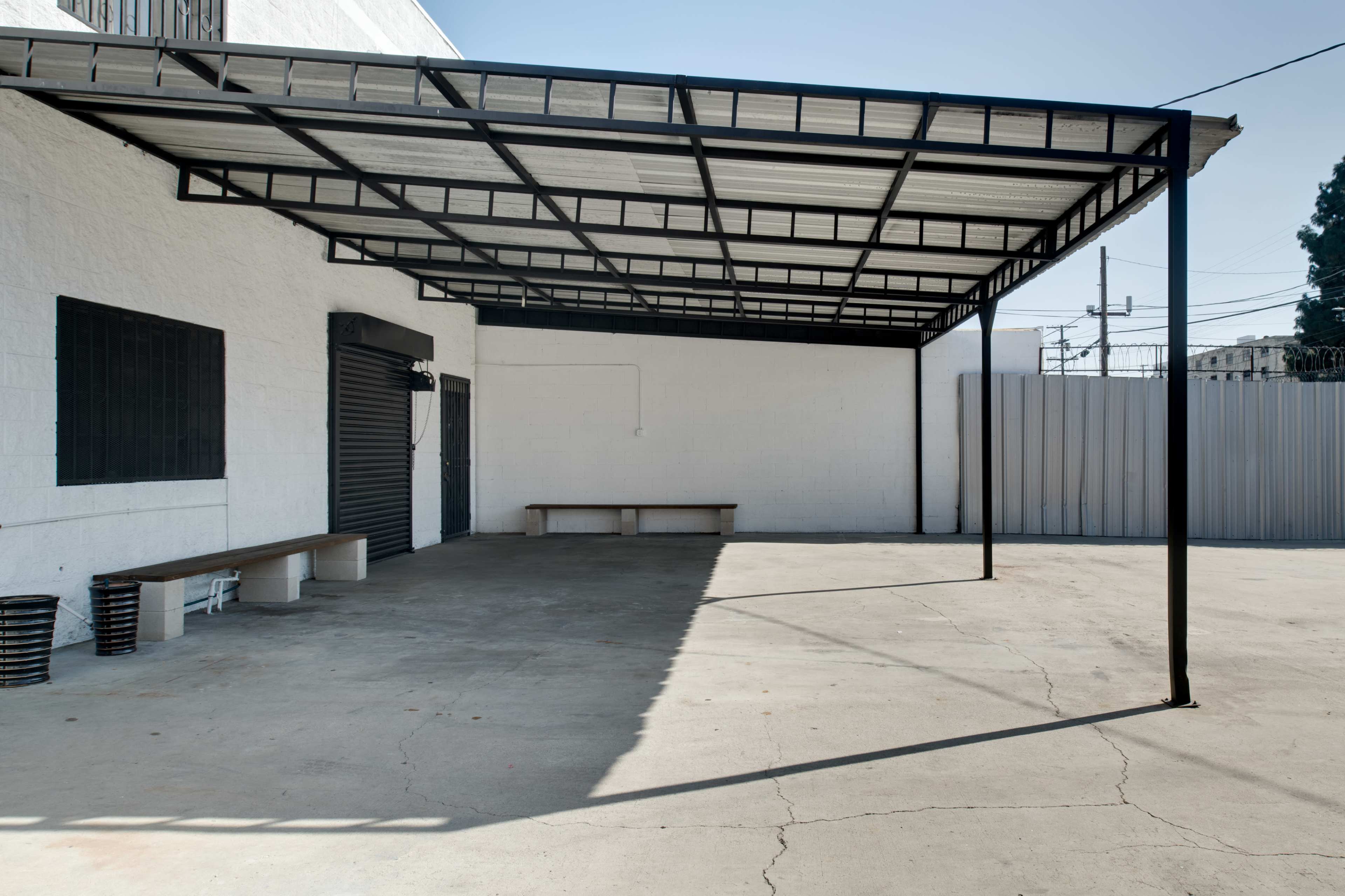 A large, open, shaded area with a metal frame structure attached to a white wall, featuring a concrete floor and a wooden bench along one side.