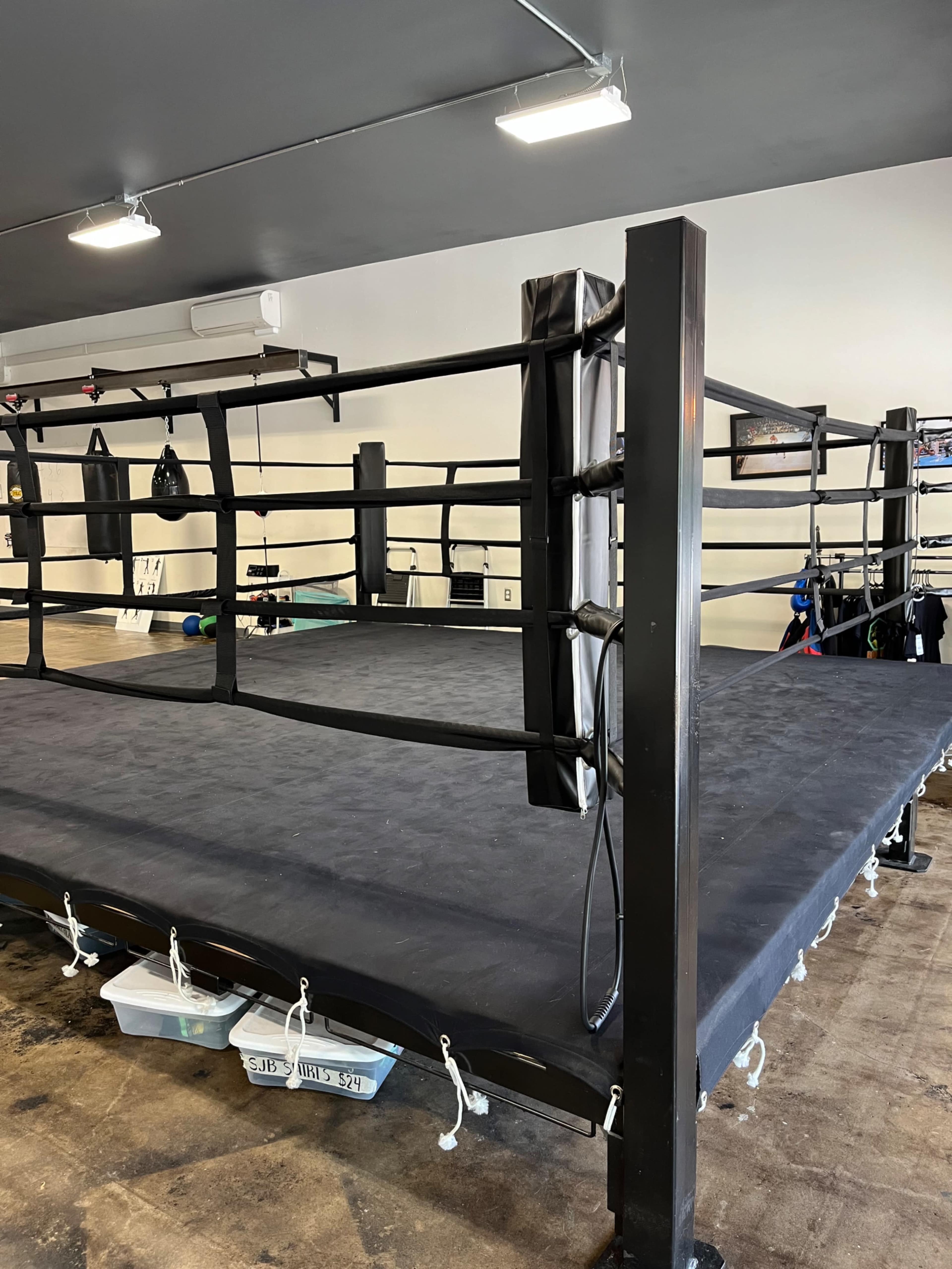 A boxing ring in a gym with black padding and equipment stored around the edges.