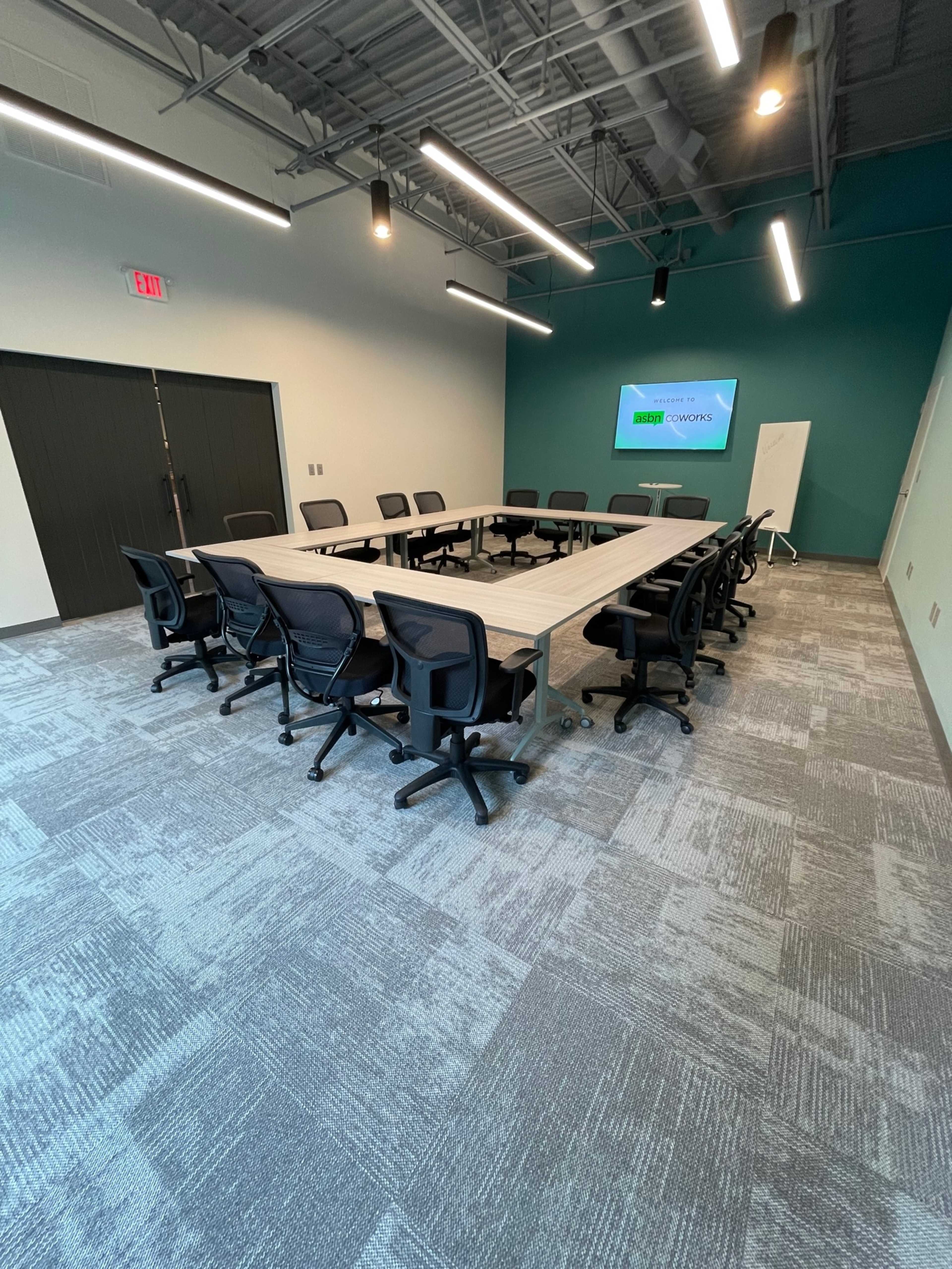 A large conference room features a long, rectangular table surrounded by ergonomic black chairs, with a screen mounted on the wall and soft lighting overhead.