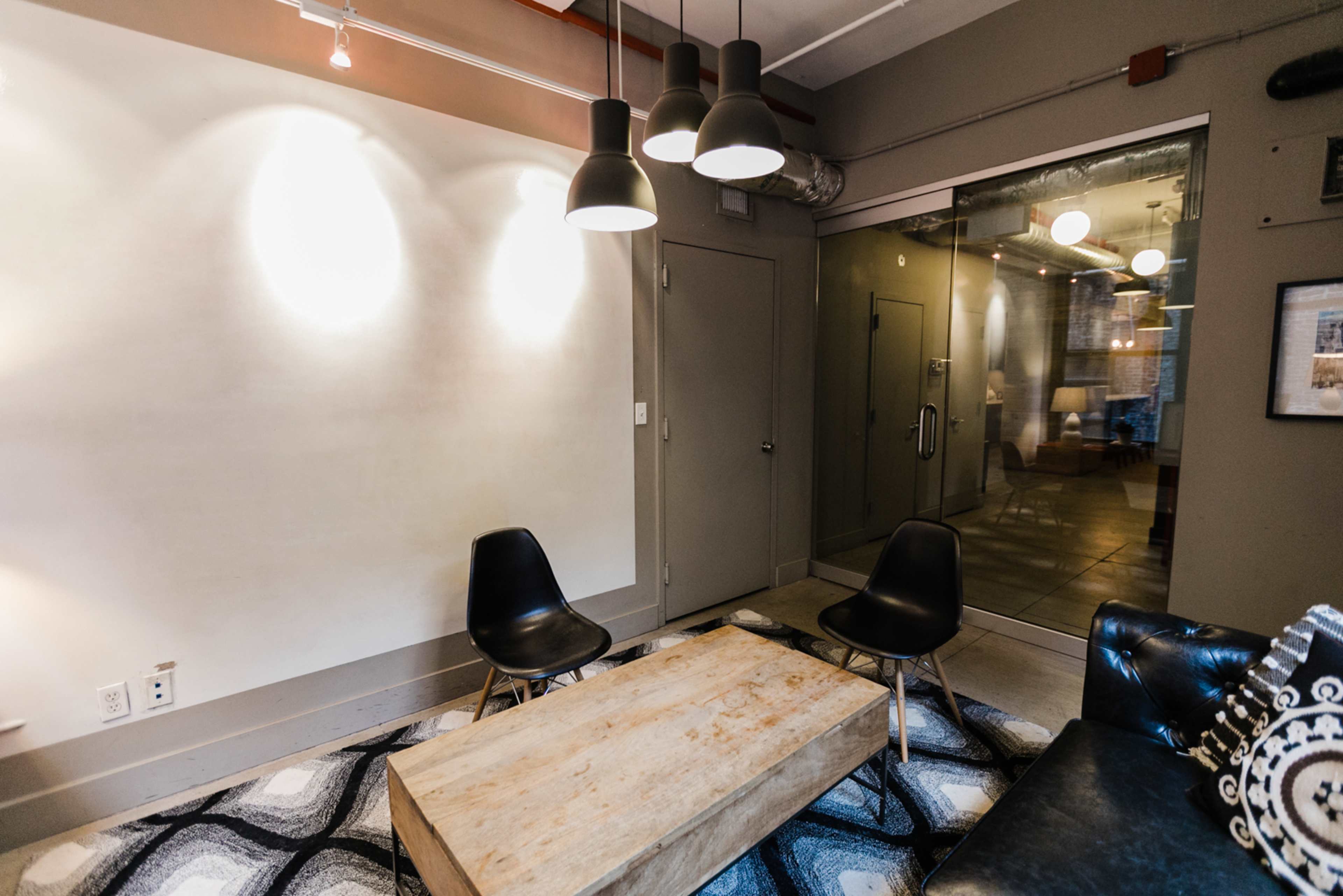 The image shows a modern lounge area with two black chairs and a wooden coffee table, illuminated by pendant lights, next to a glass door leading to another room.