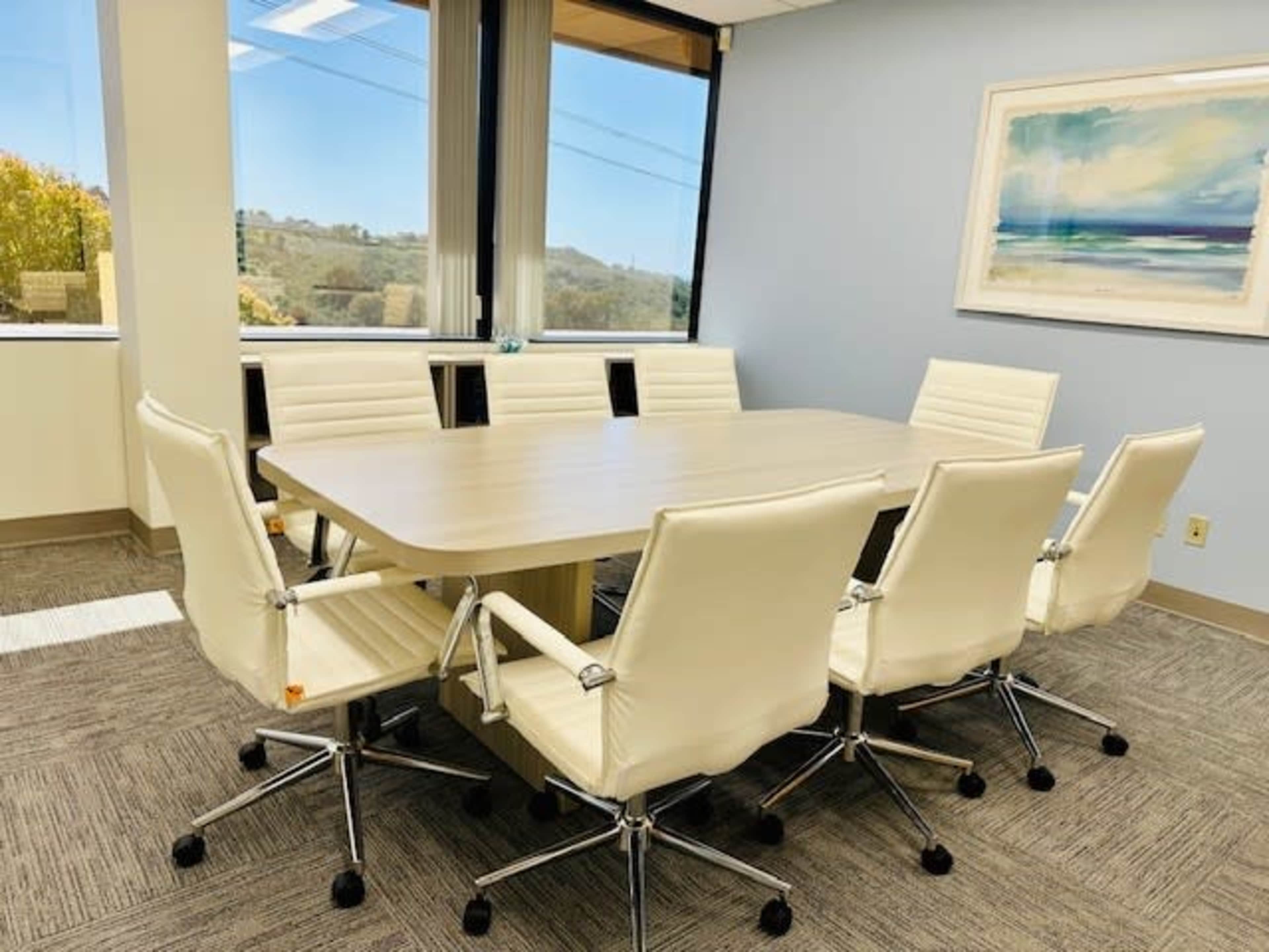 A modern conference room features a large, rectangular table surrounded by eight white upholstered chairs, with large windows offering a view of greenery outside.