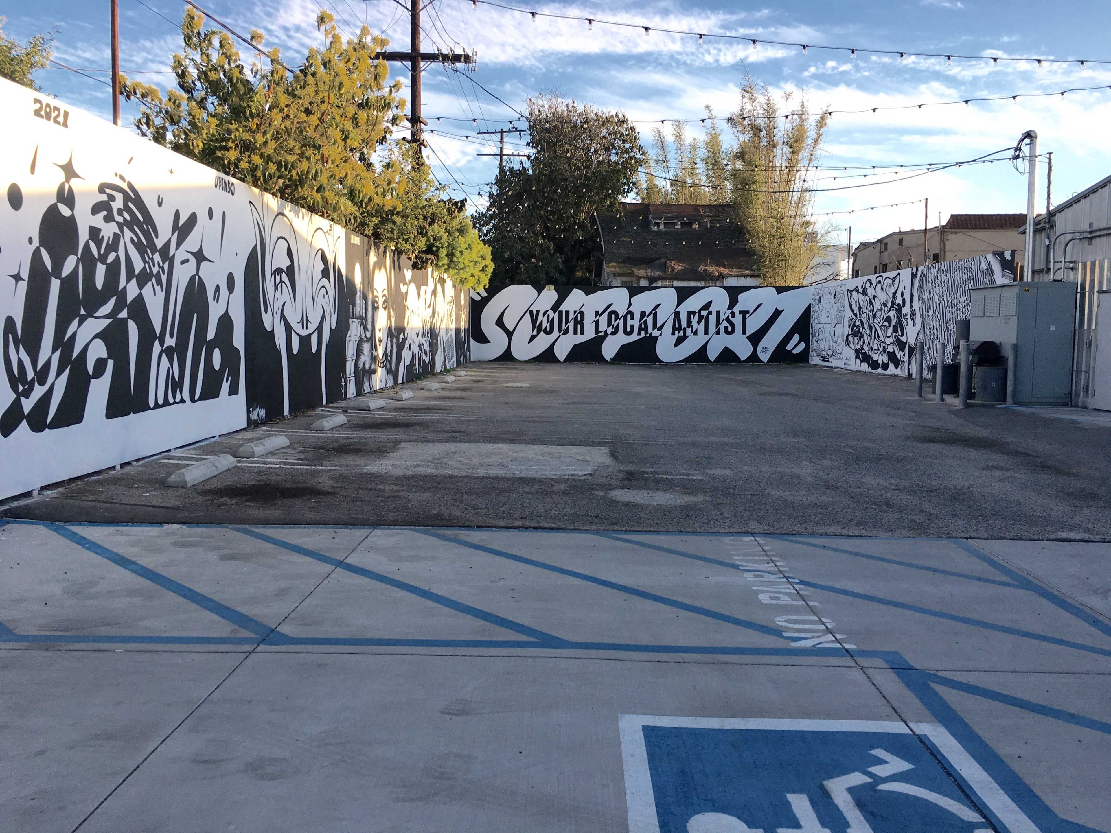The image shows a graffiti-covered wall in an open area with a parking lot marked for accessible parking.