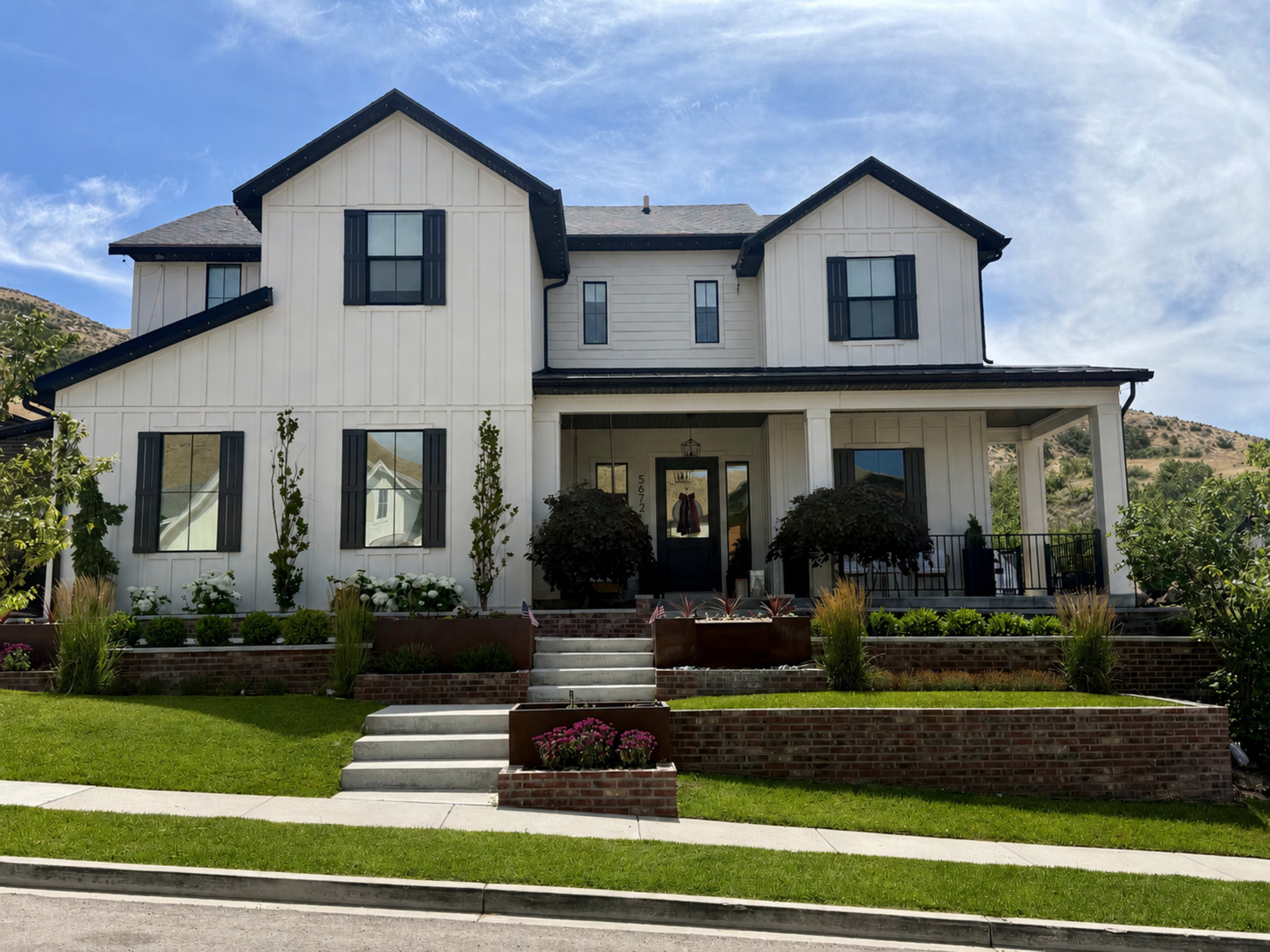 The image shows a two-story white farmhouse-style house with a large front porch, manicured lawns, and a landscaped garden.