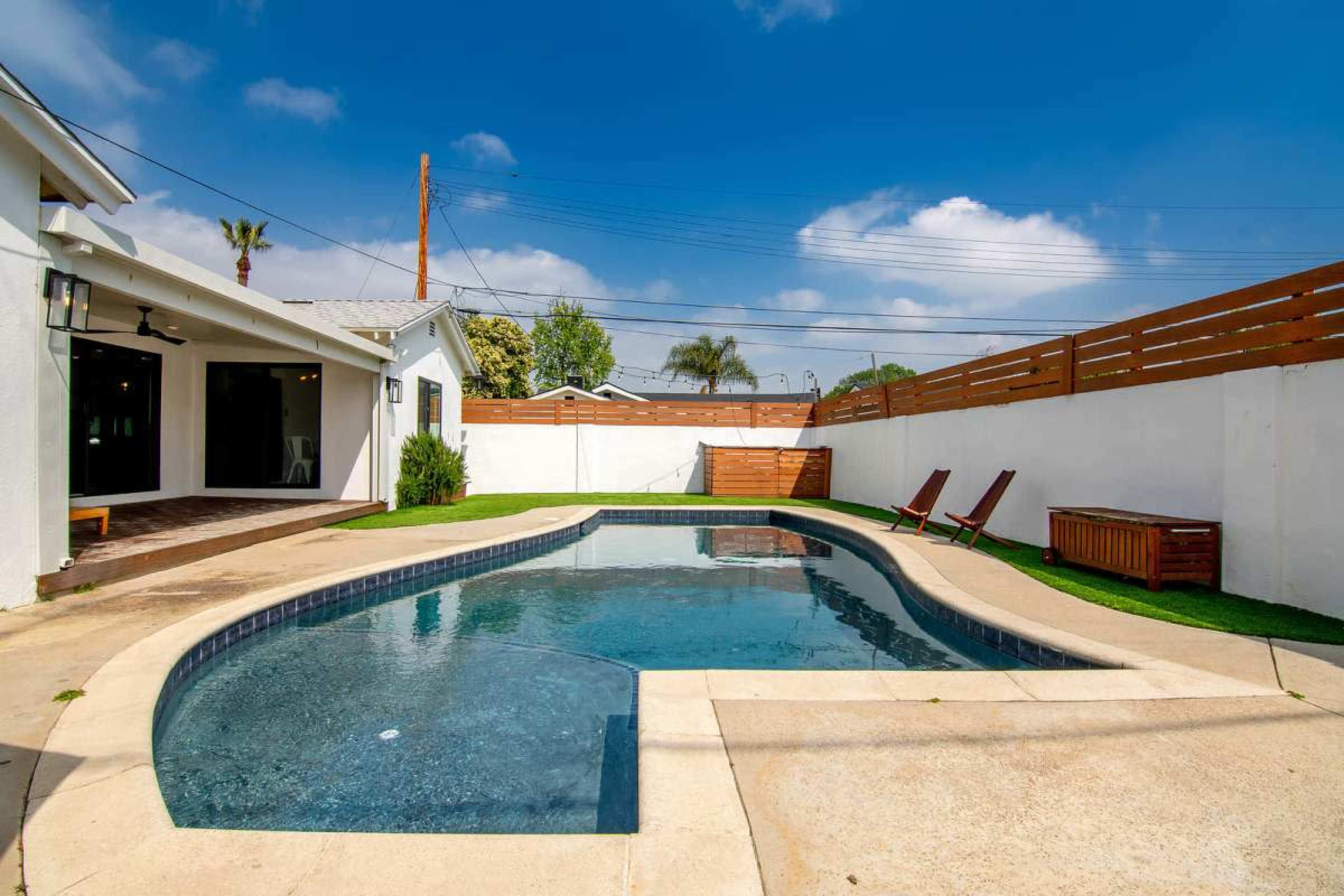 The image shows a swimming pool surrounded by a patio, with two lounge chairs and a wooden storage box beside a white fence.