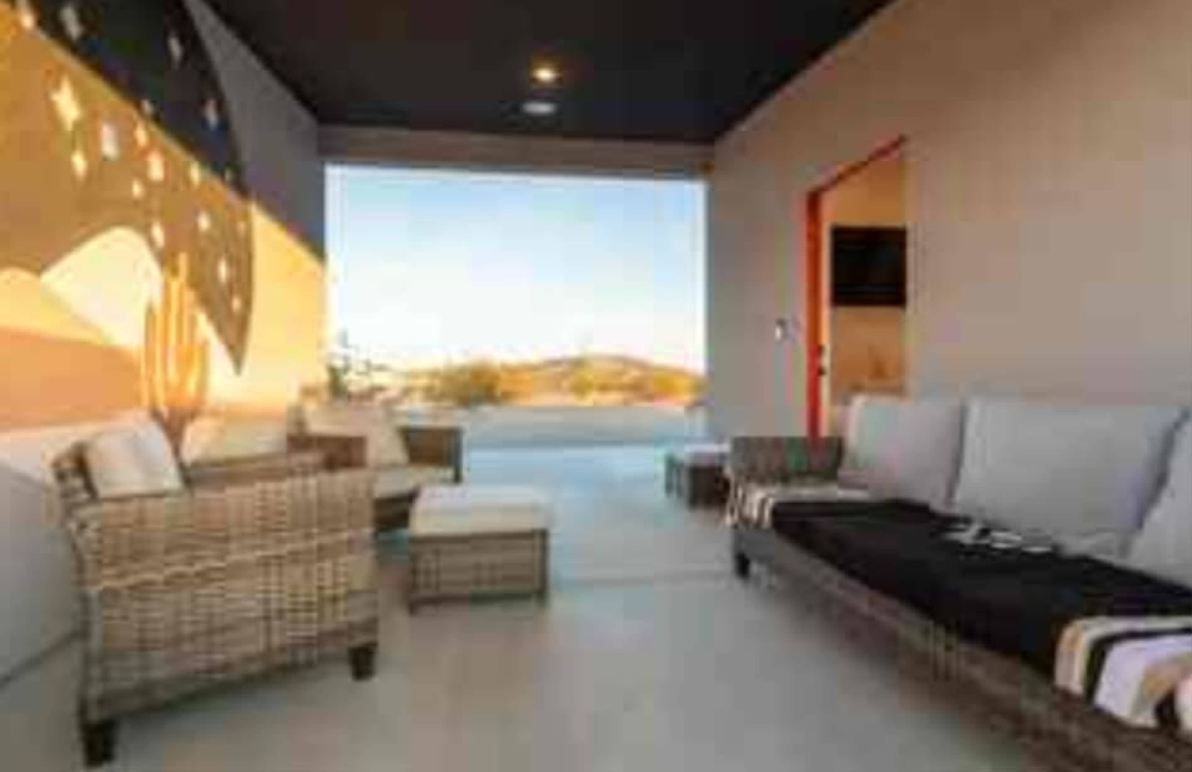 The image shows a modern outdoor lounge area with woven furniture and a large mural on the wall, framed by a view of a desert landscape.
