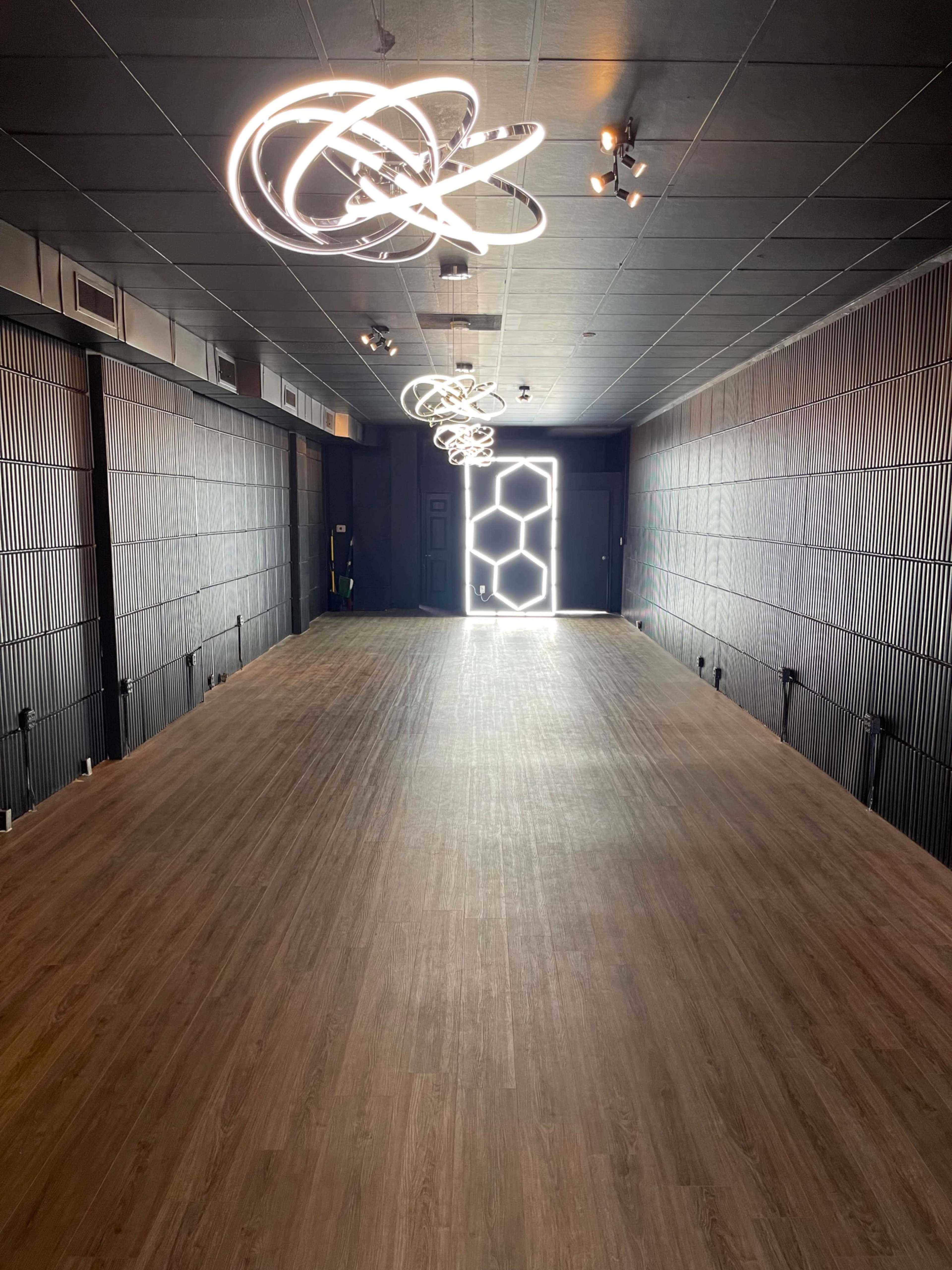 A spacious interior room with wooden flooring, illuminated by modern light fixtures and featuring a hexagonal doorway at the end.