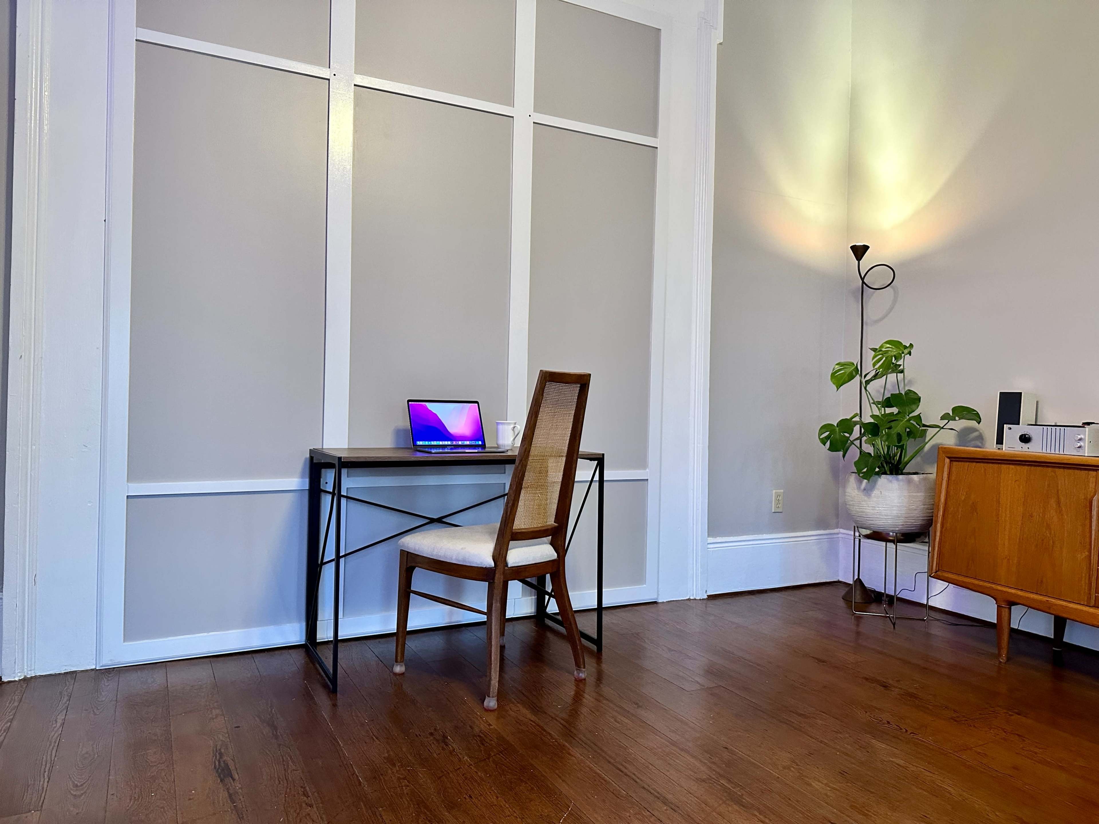 A simple desk with a laptop is positioned near a wall, accompanied by a chair and a potted plant in a well-lit room.