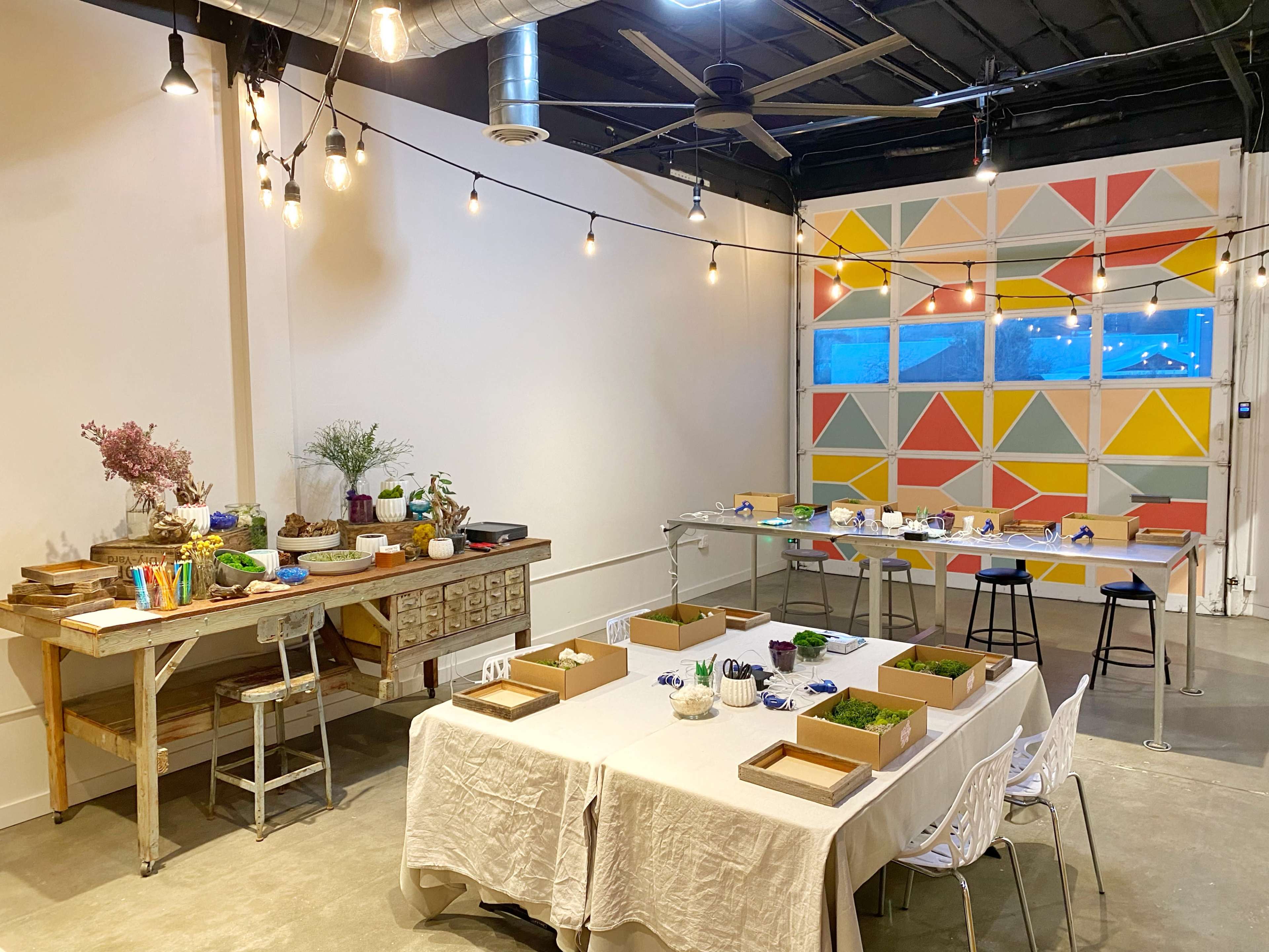 A well-lit creative workshop space with a large table covered in a white cloth, various art supplies organized on a wooden shelf, and colorful geometric wall decor.