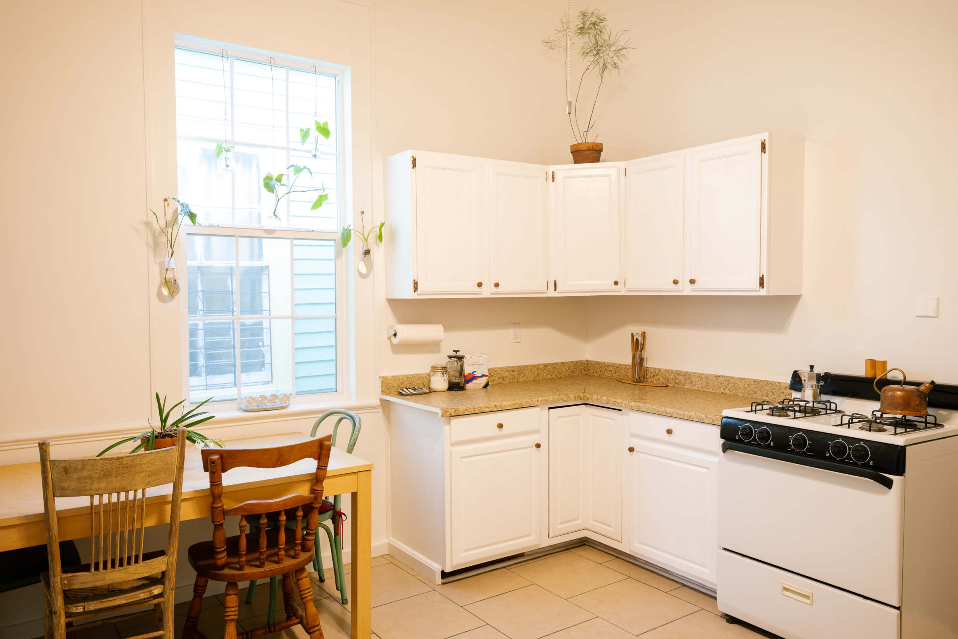 The image shows a small kitchen with white cabinets, a stove, and a table with chairs near a window.