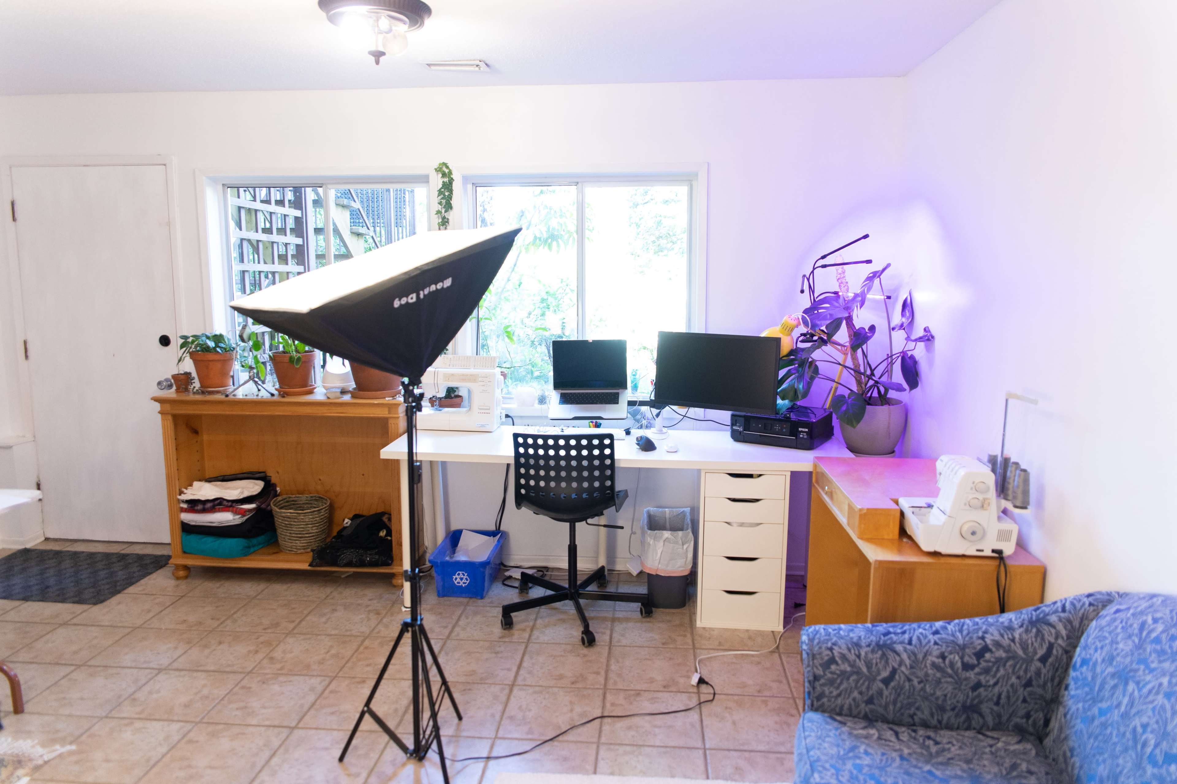 A home workspace featuring a desk with two computer monitors, a potted plant, and a photography light setup in a tiled room.
