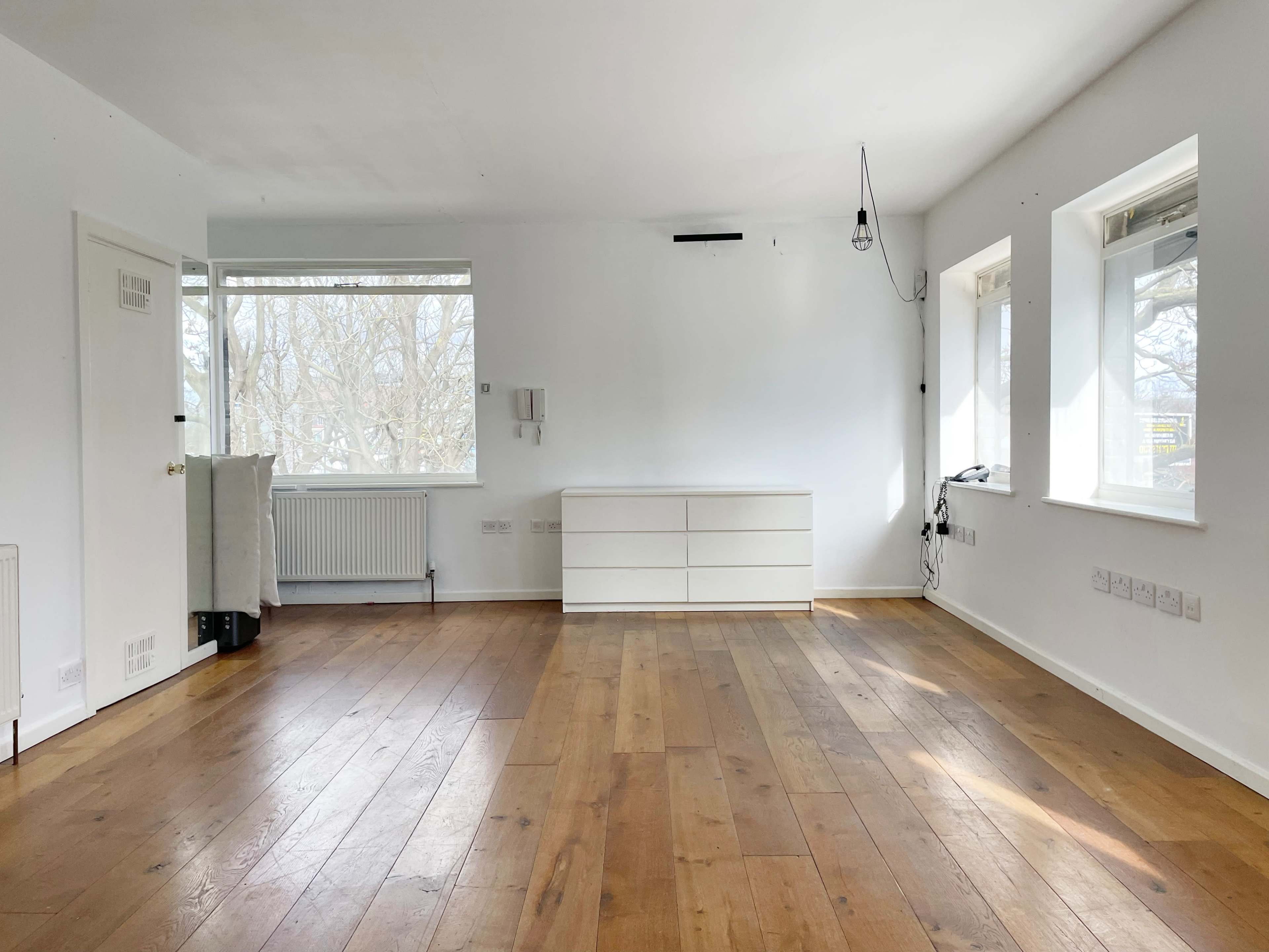 A spacious, empty room with light wood flooring and a white dresser against one wall, featuring large windows that allow natural light to fill the space.