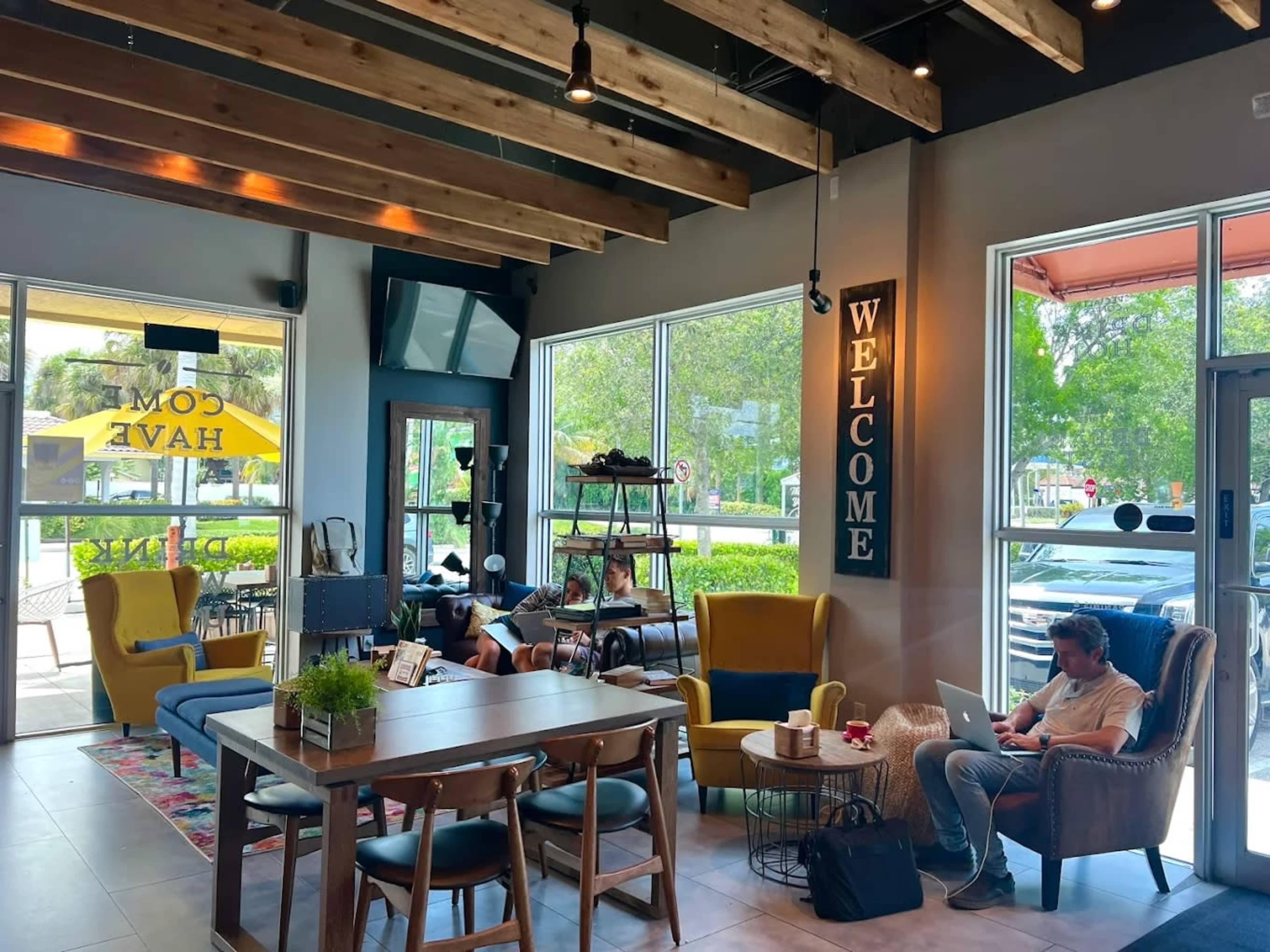 A cozy café features blue and yellow seating, large windows allowing natural light, and a sign that reads "WELCOME."