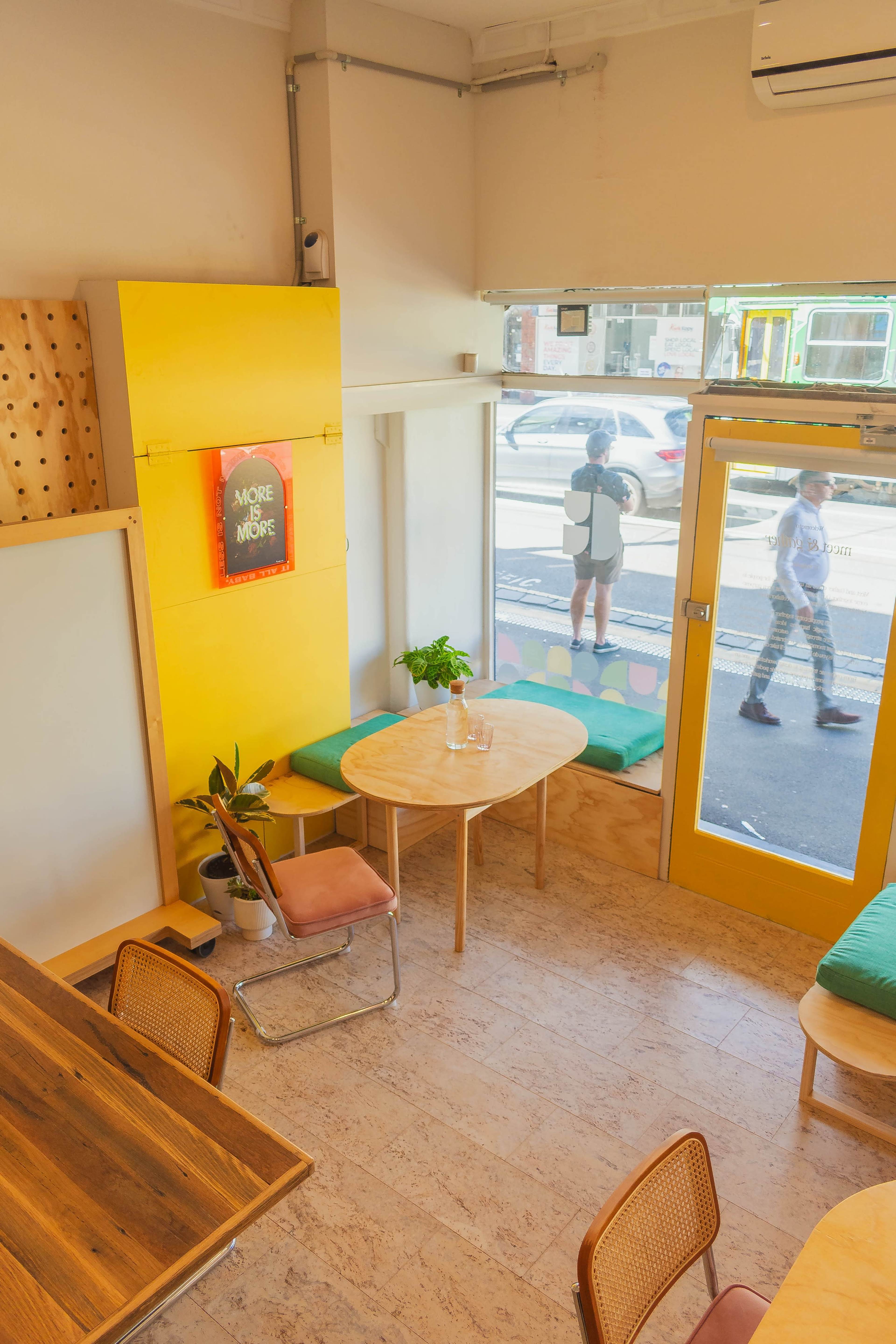 A brightly lit café features yellow walls, wooden tables, and a view of pedestrians outside through large windows.