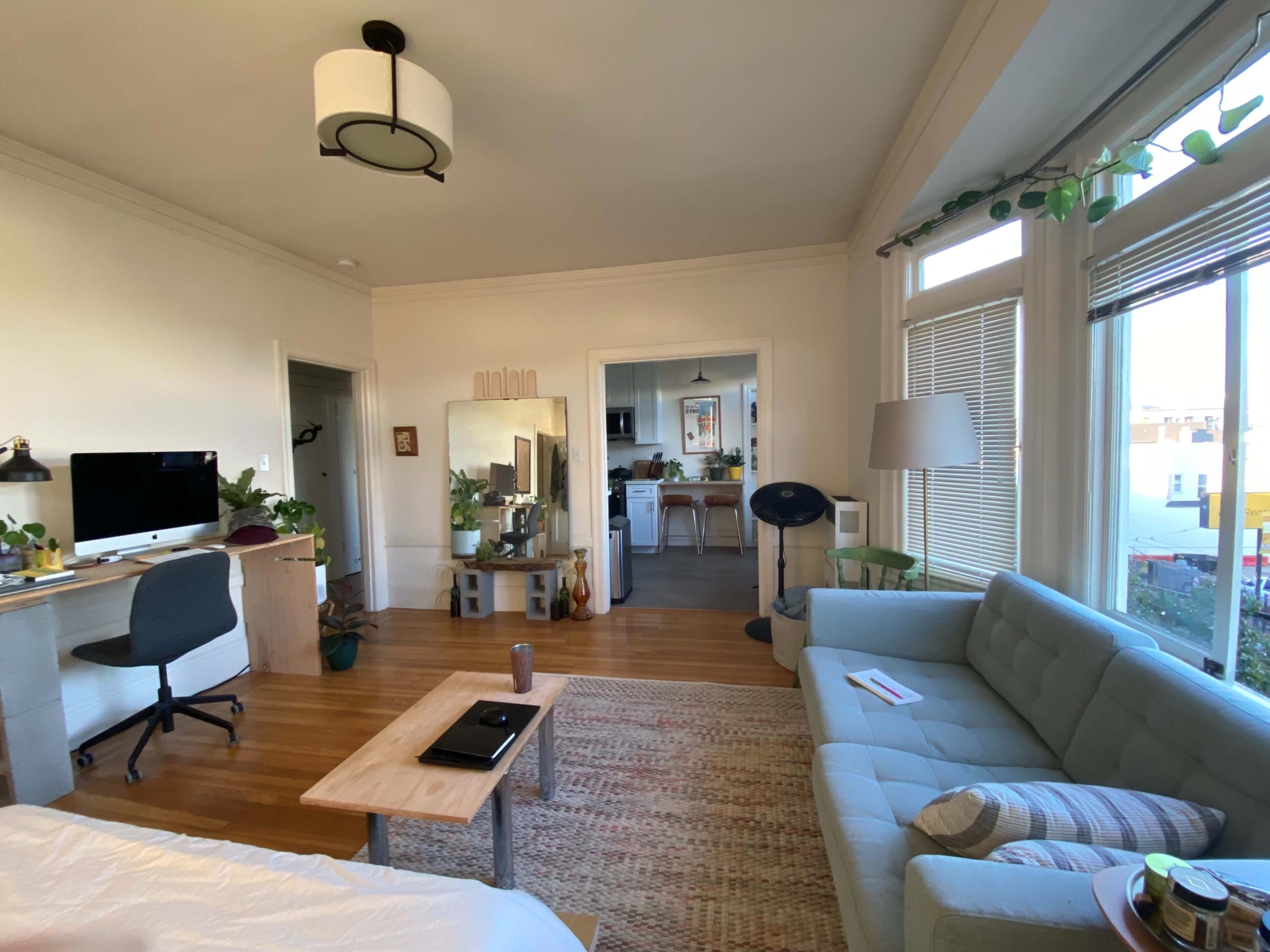 A bright living space featuring a light-colored sofa, a wooden coffee table, a desk with a computer, and an open kitchen area in the background.