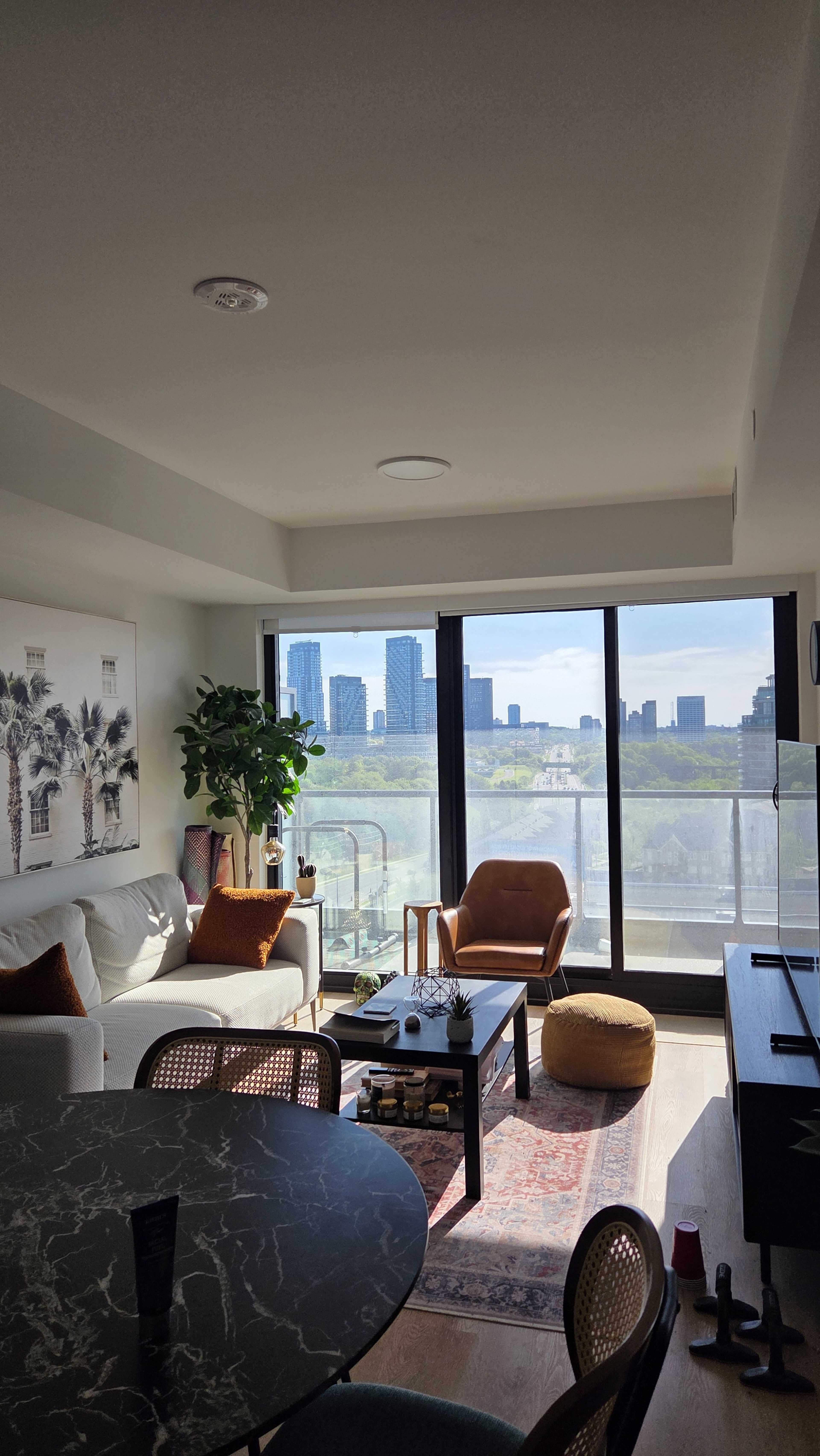 The image shows a modern living room with a view of city skyscrapers through large glass doors, featuring a cozy seating area and a round dining table.