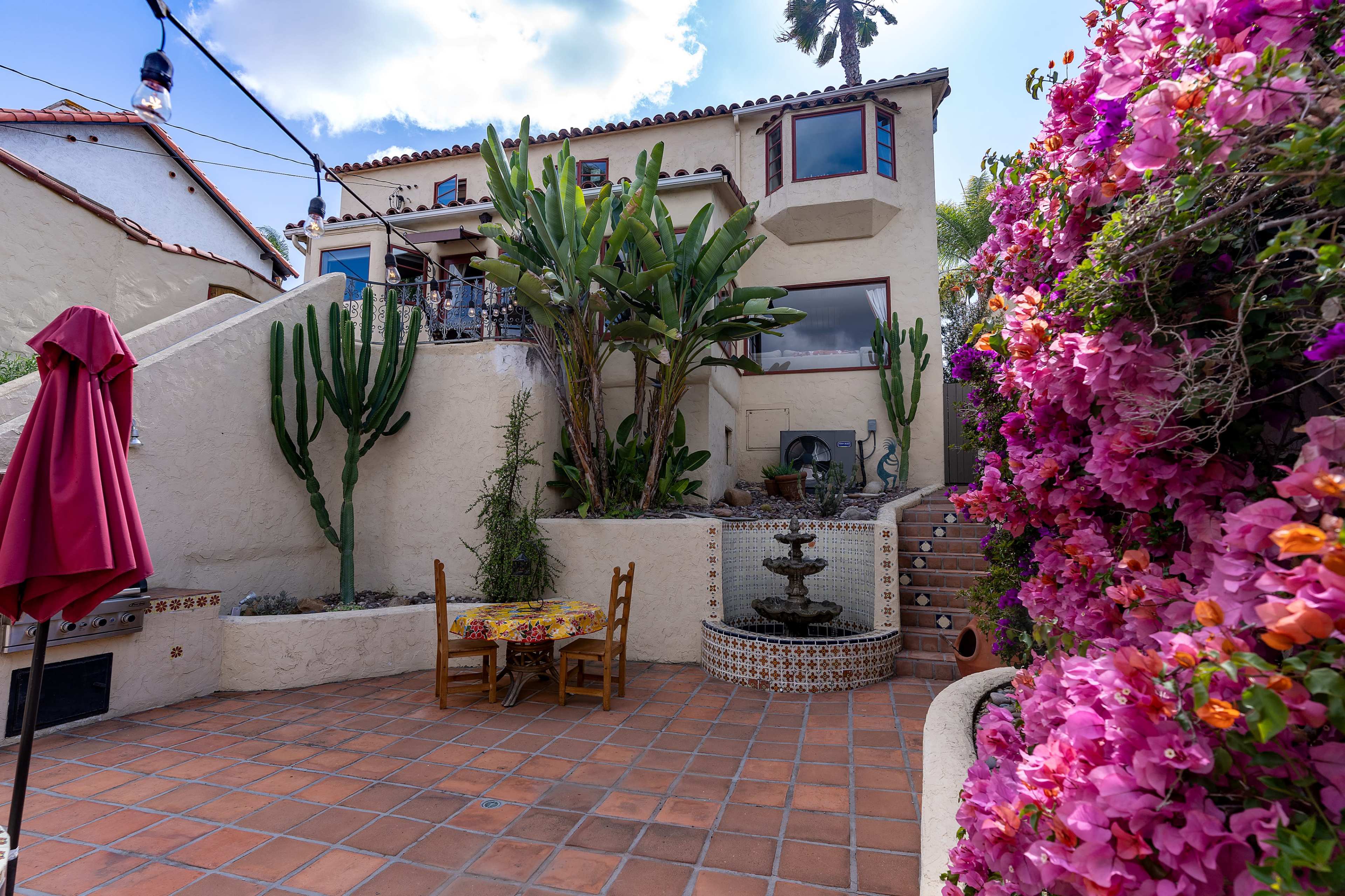 A patio area with a tiled fountain, surrounded by vibrant flowers and cacti, adjacent to a multi-story house.