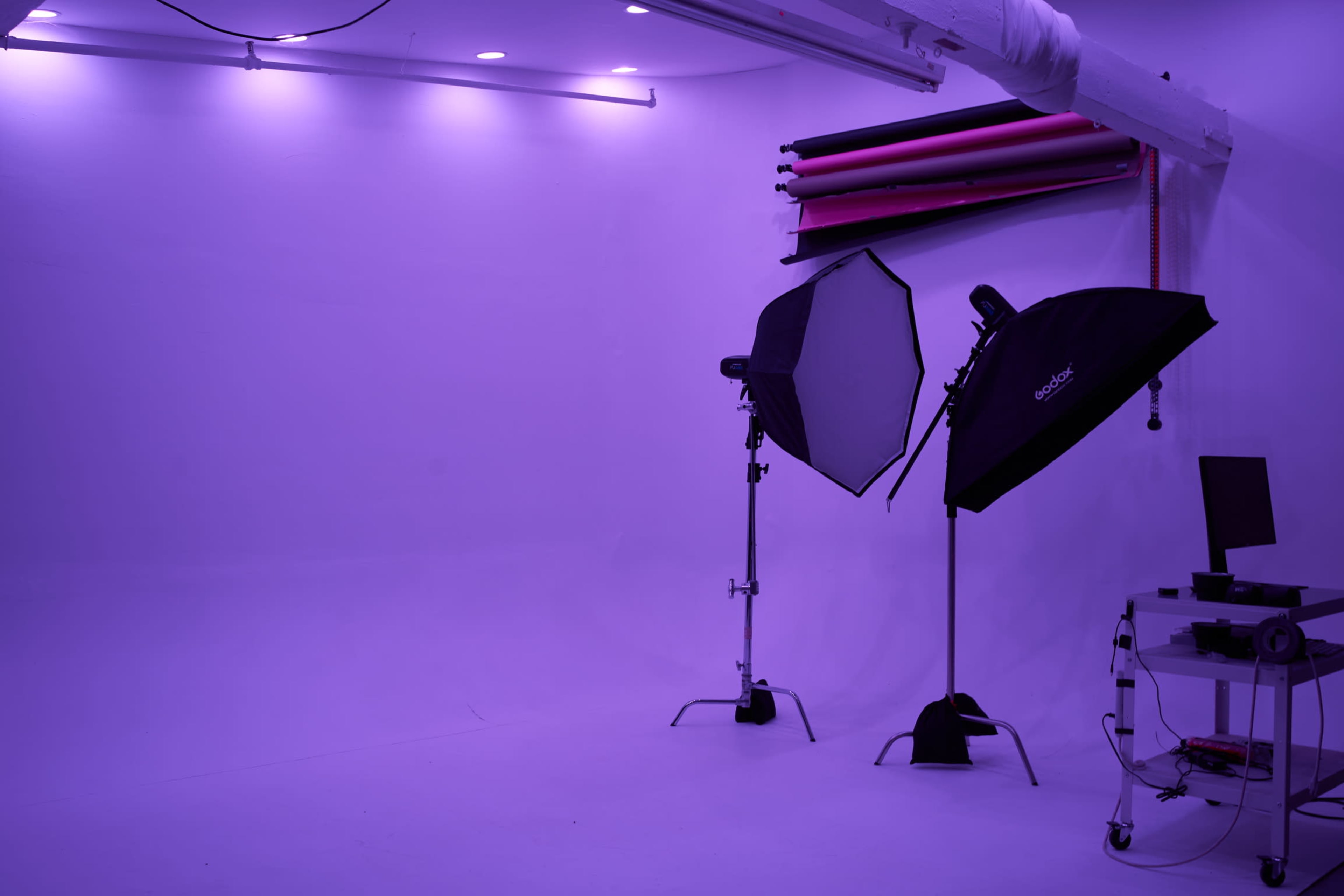 The image shows a photography studio with two light setups and a computer station against a purple backdrop.