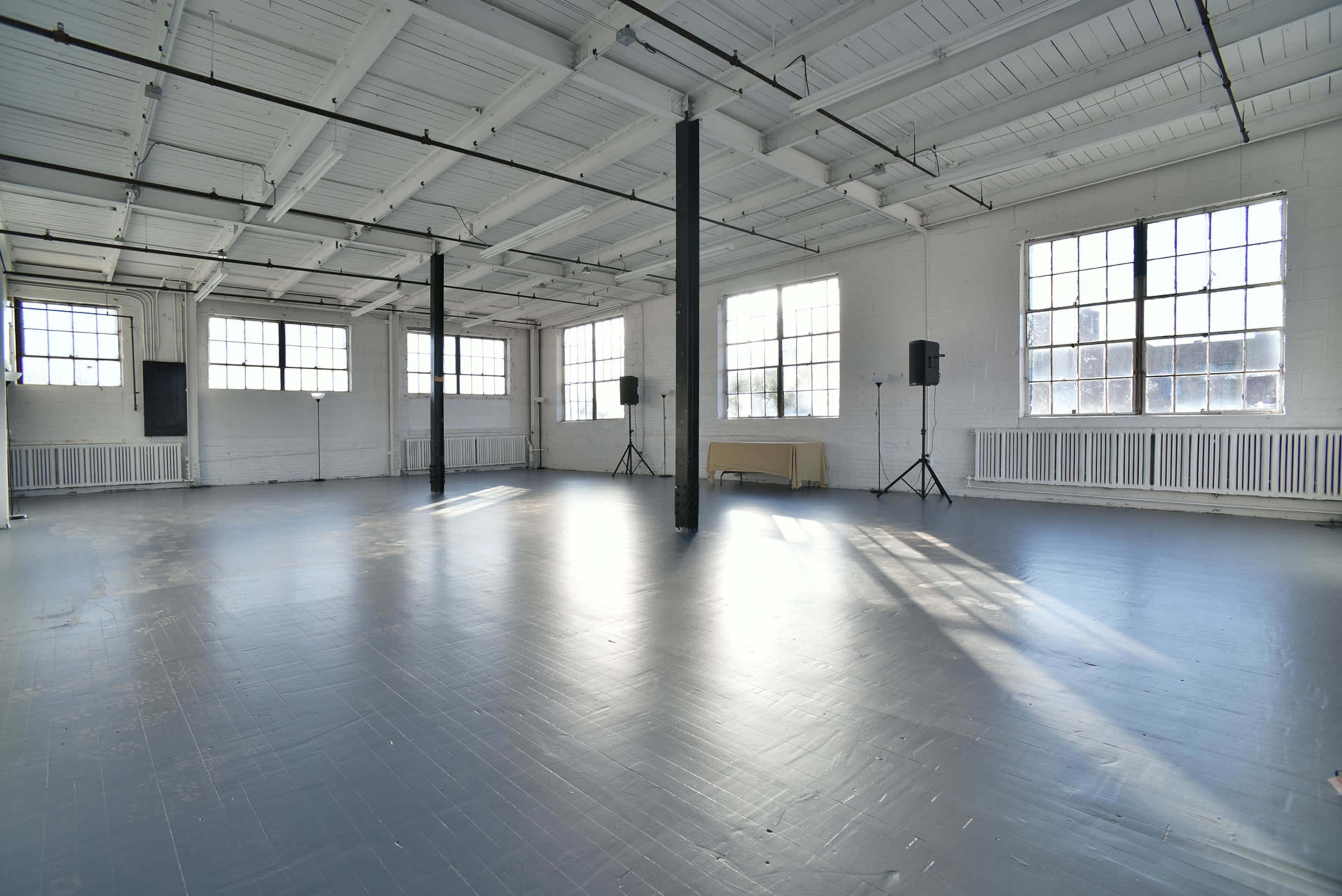 A spacious, well-lit interior with large windows and polished gray flooring, featuring two speakers on stands and a table against the wall.