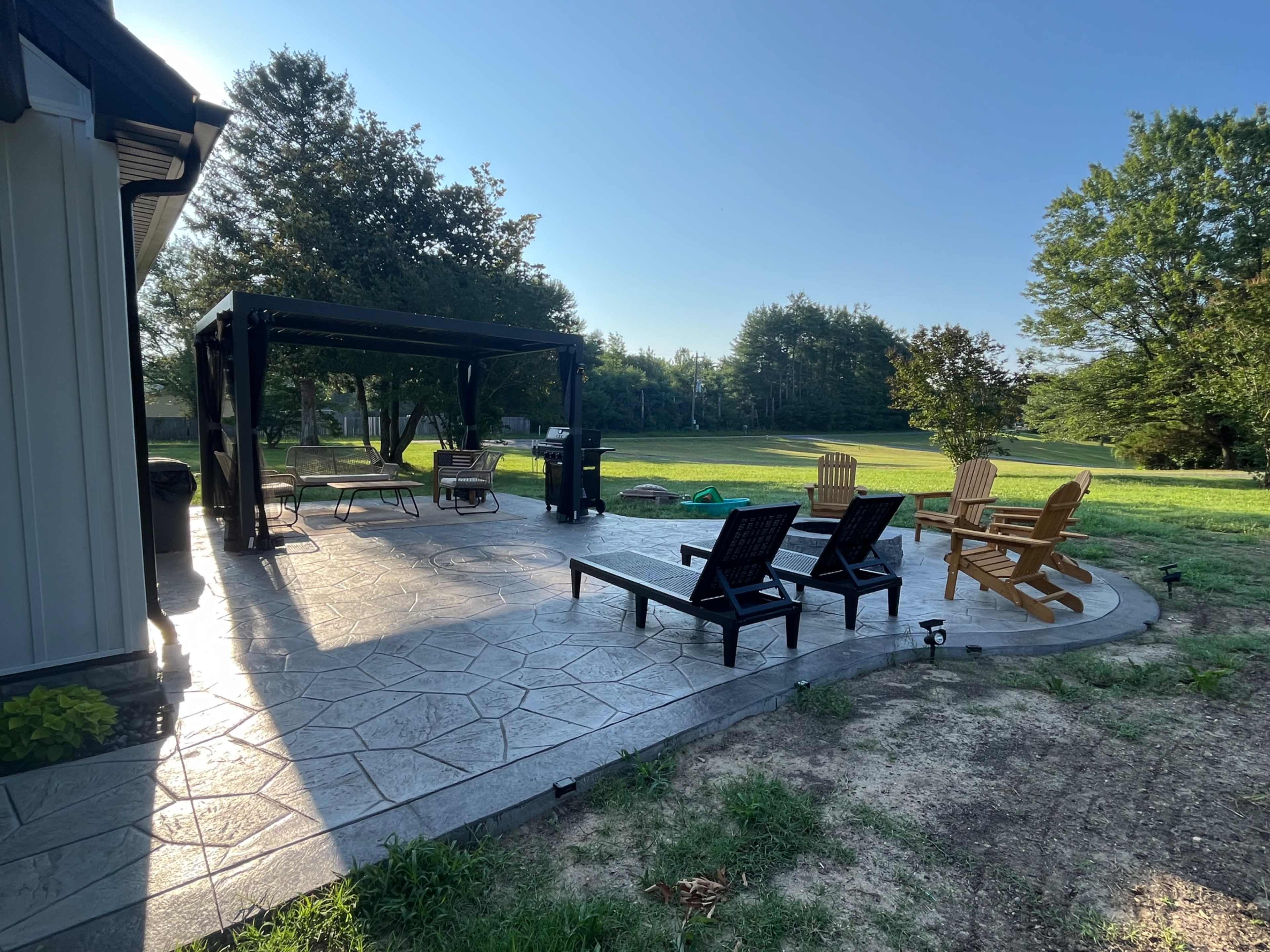 The image shows a patio area with several chairs, a pergola structure, and a lawn in the background under clear blue skies.