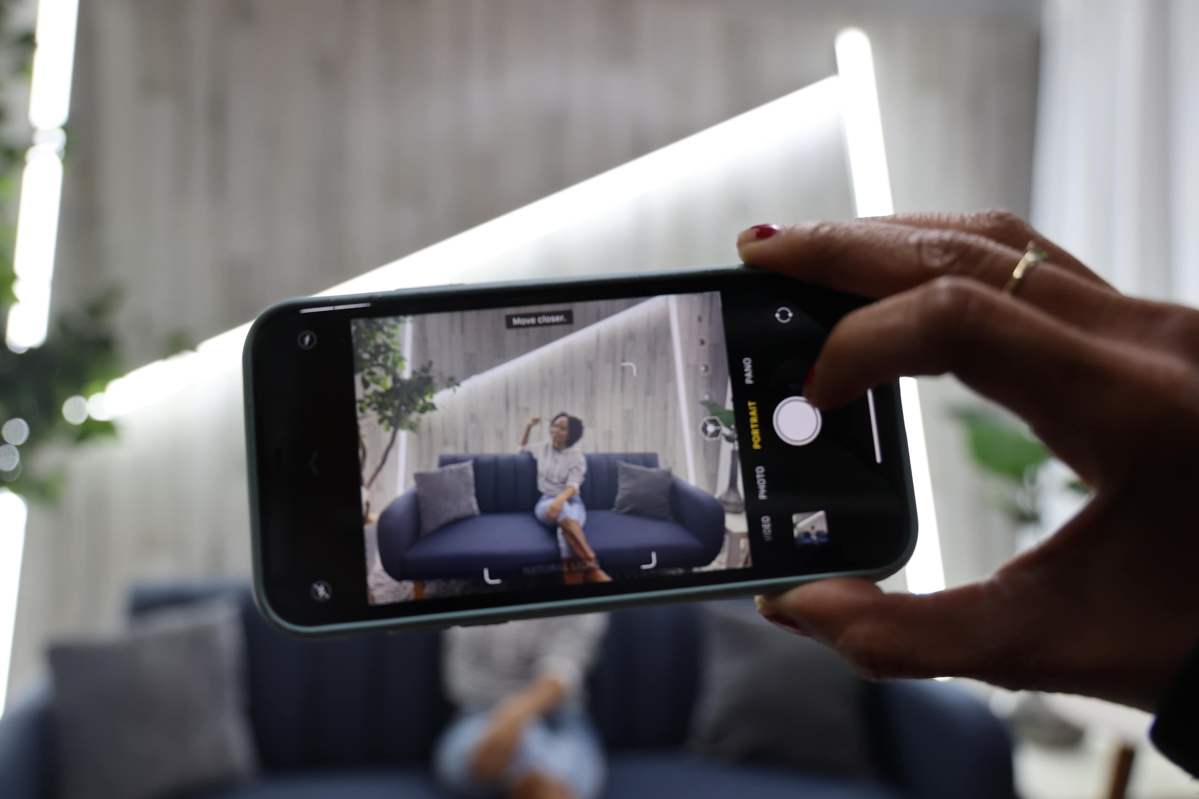 A person holds a smartphone, capturing an image of another individual sitting on a couch against a backdrop with decorative lighting.