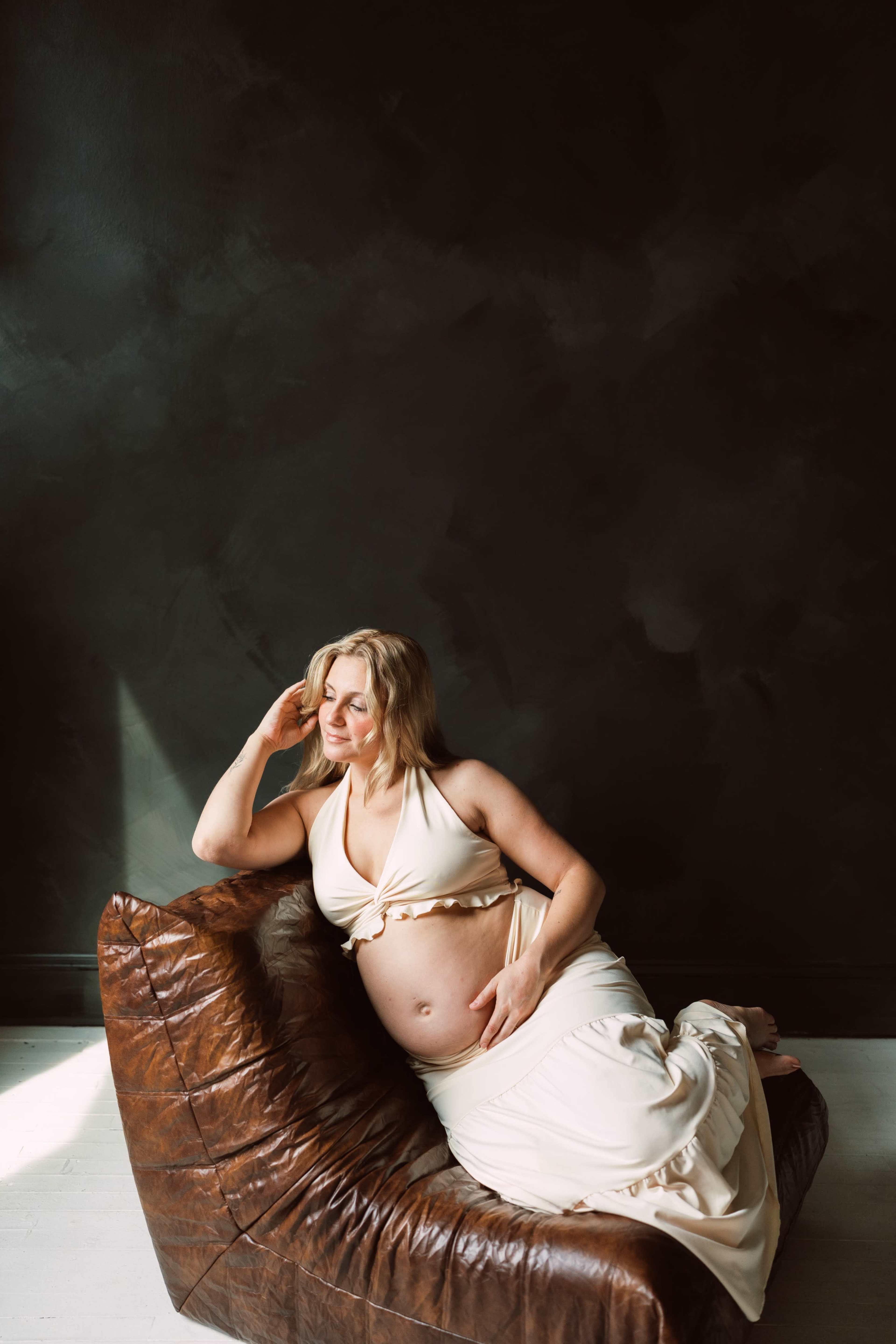 A pregnant woman in a flowing dress sits on a brown leather couch against a dark wall.