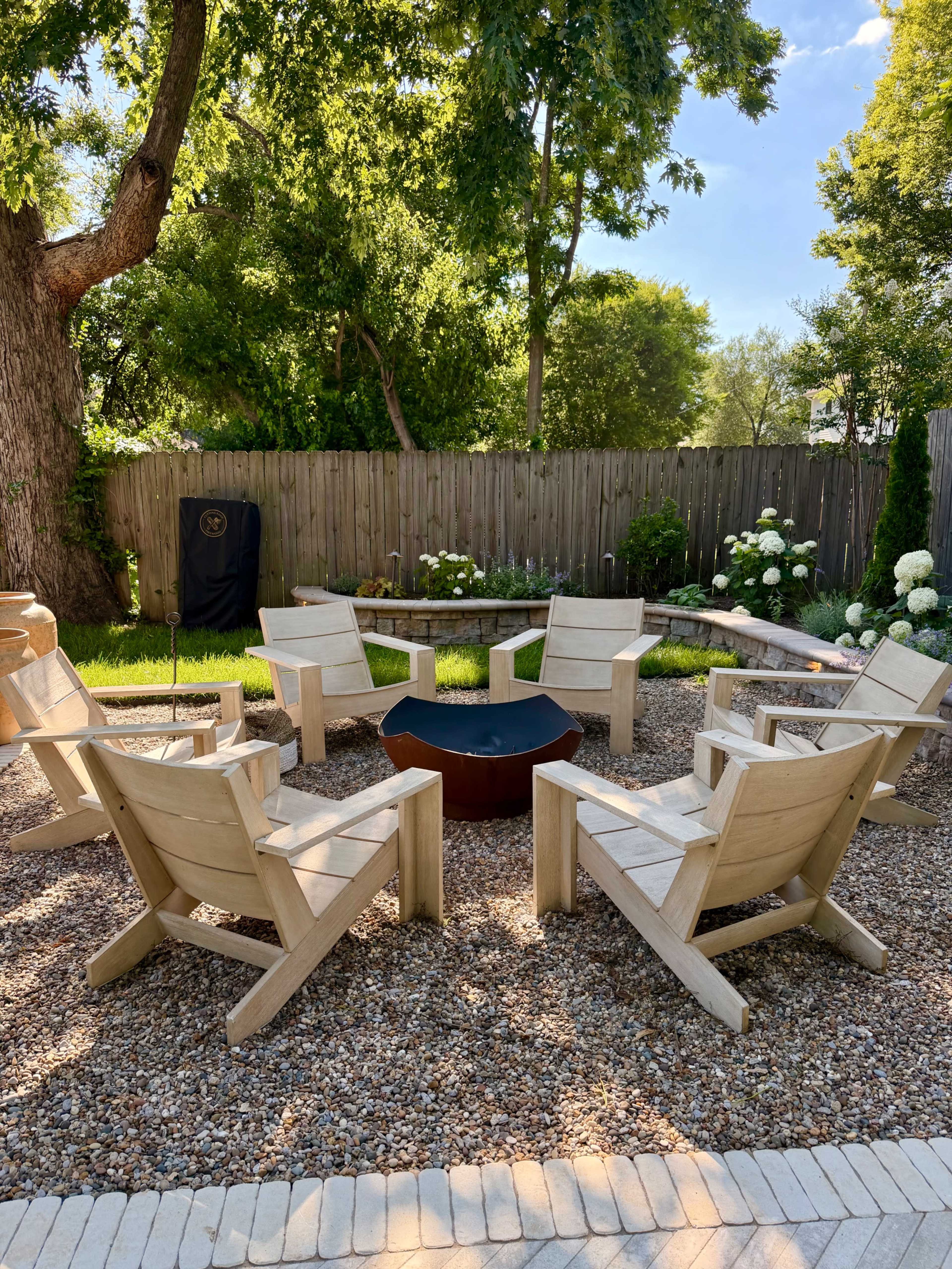 The image shows a circular seating area with wooden chairs surrounding a fire pit, set in a landscaped backyard with greenery and flower beds.