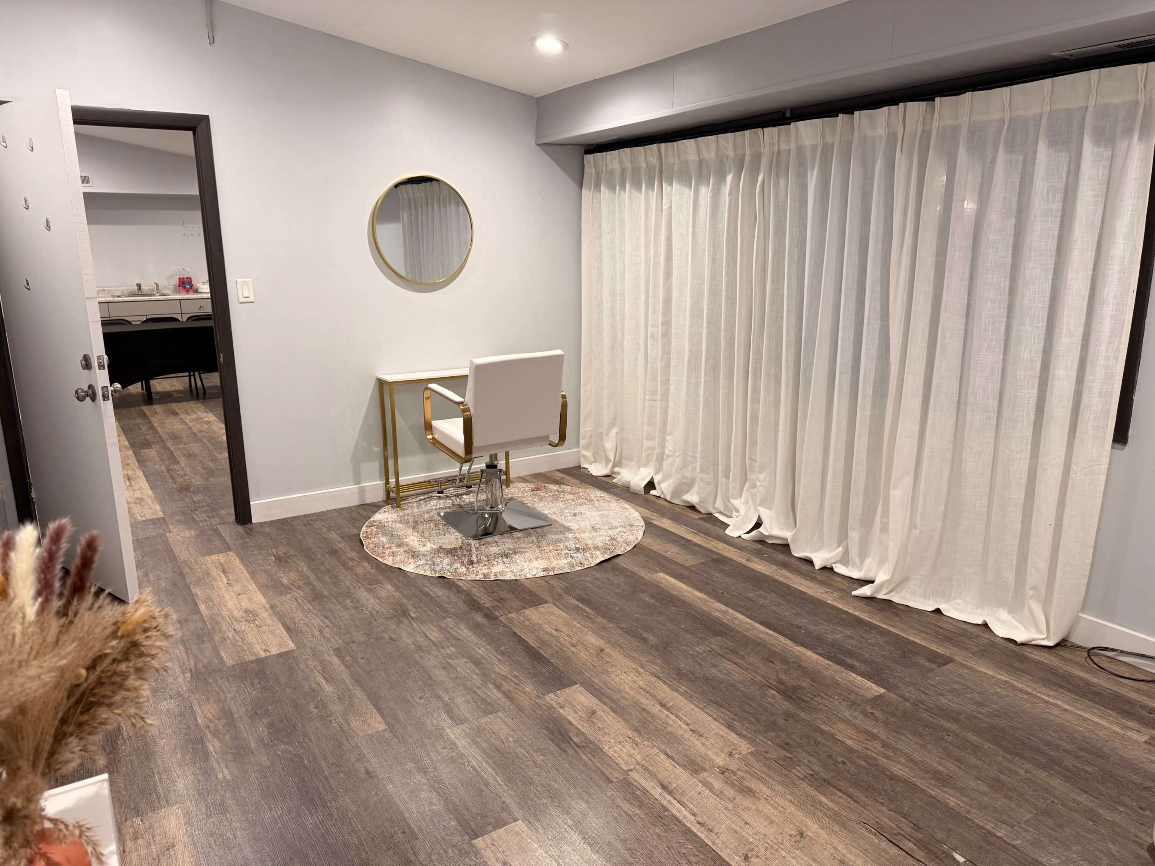 The image shows a modern interior space featuring a small desk with a white chair and a round rug, surrounded by light-colored walls and a large window covered with sheer curtains.