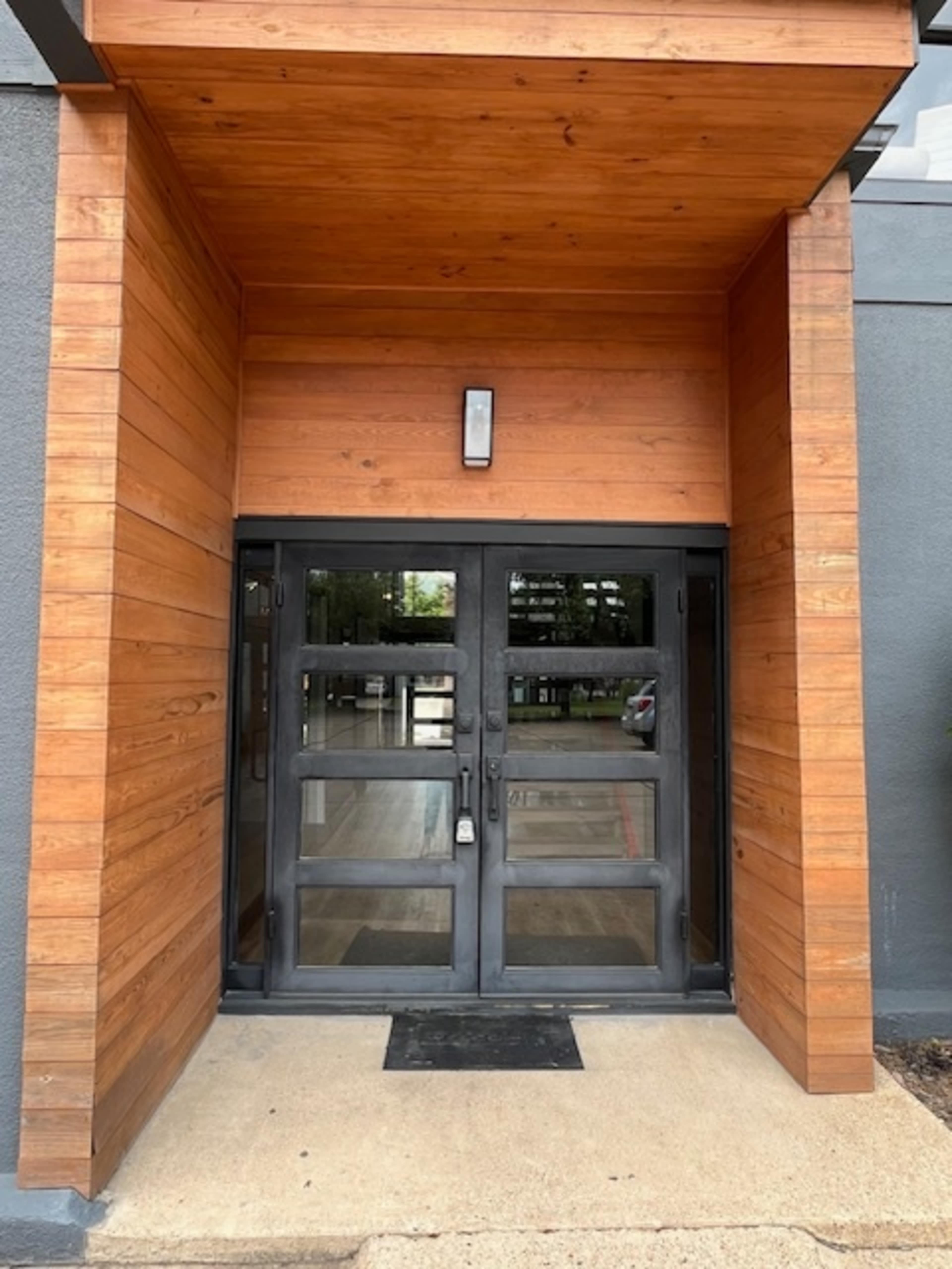 The image shows a set of double glass doors framed by wooden panels and a flat wooden overhang, leading into a modern building entrance.