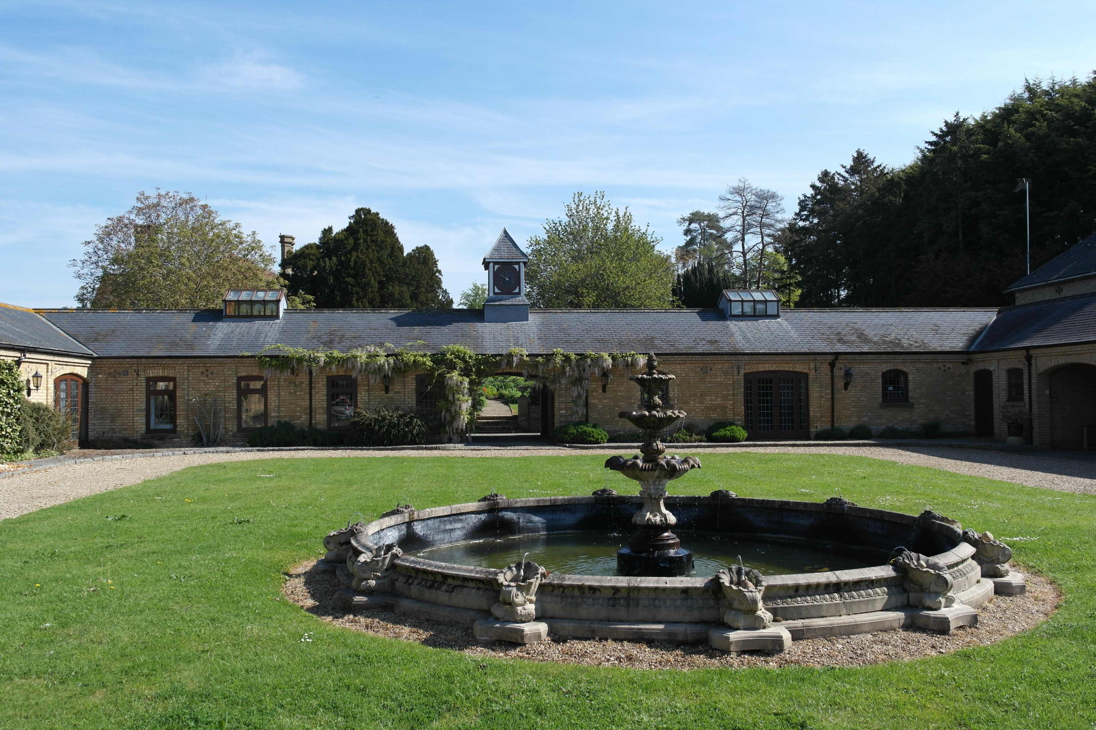 A large stone fountain sits in a grassy courtyard surrounded by buildings with windows and a clock tower in the background.