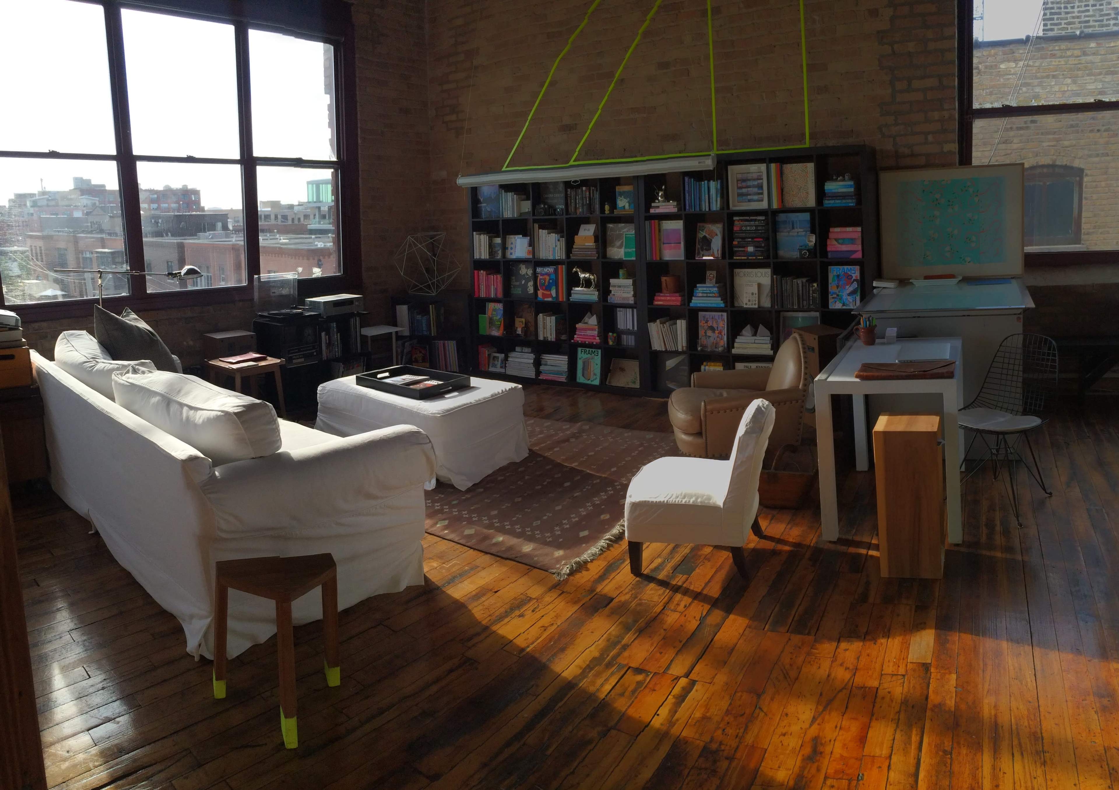 A spacious, sunlit living room with large windows, wooden floors, a white sofa, a bookshelf filled with books, and a simple desk and chair arrangement.