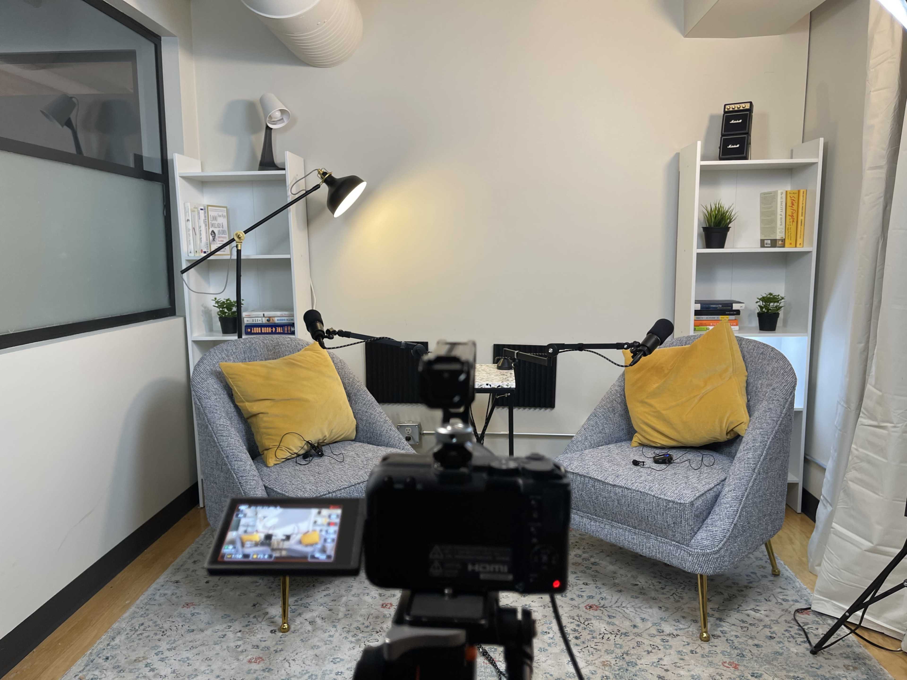 The image shows a podcast recording setup featuring two gray chairs with yellow cushions, a microphone on each chair, and a camera positioned in front, surrounded by shelves containing books and decorative plants.