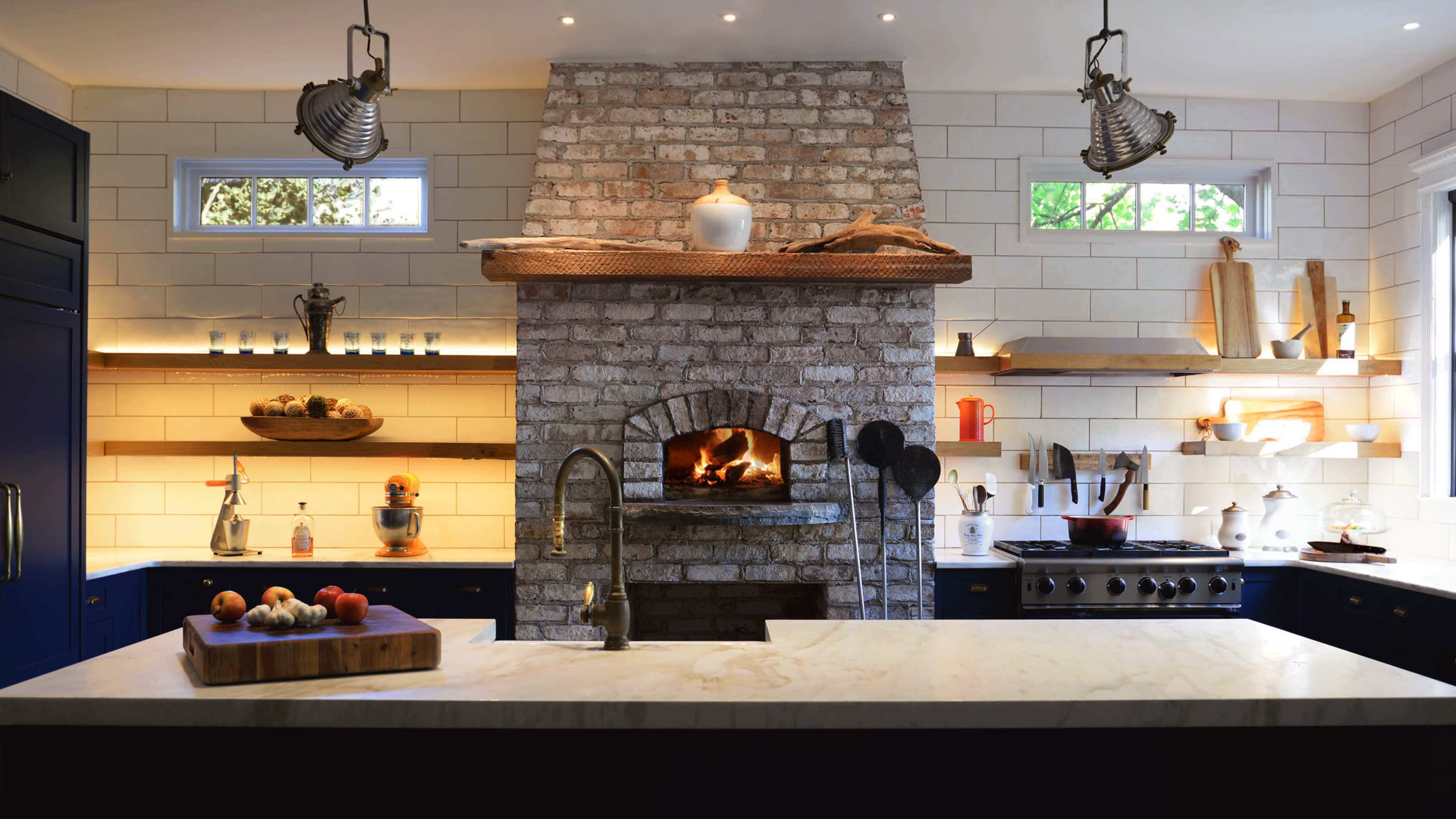 A modern kitchen features a large brick fireplace with a wooden mantel, stainless steel appliances, and open shelves displaying various kitchen items.