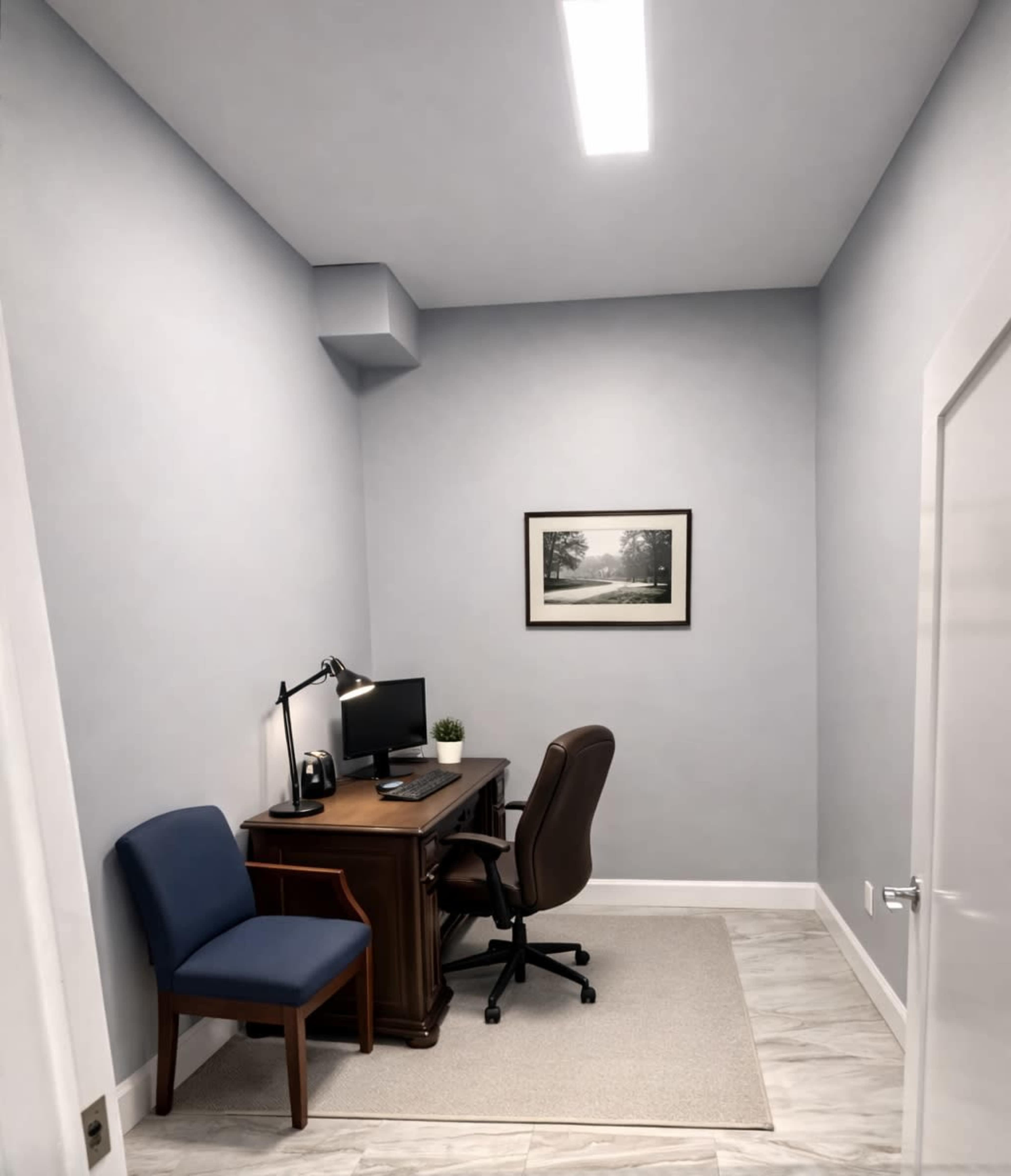 The image shows a small office space featuring a wooden desk with a computer, a task lamp, a chair, a blue upholstered chair, and a framed photograph on the wall.