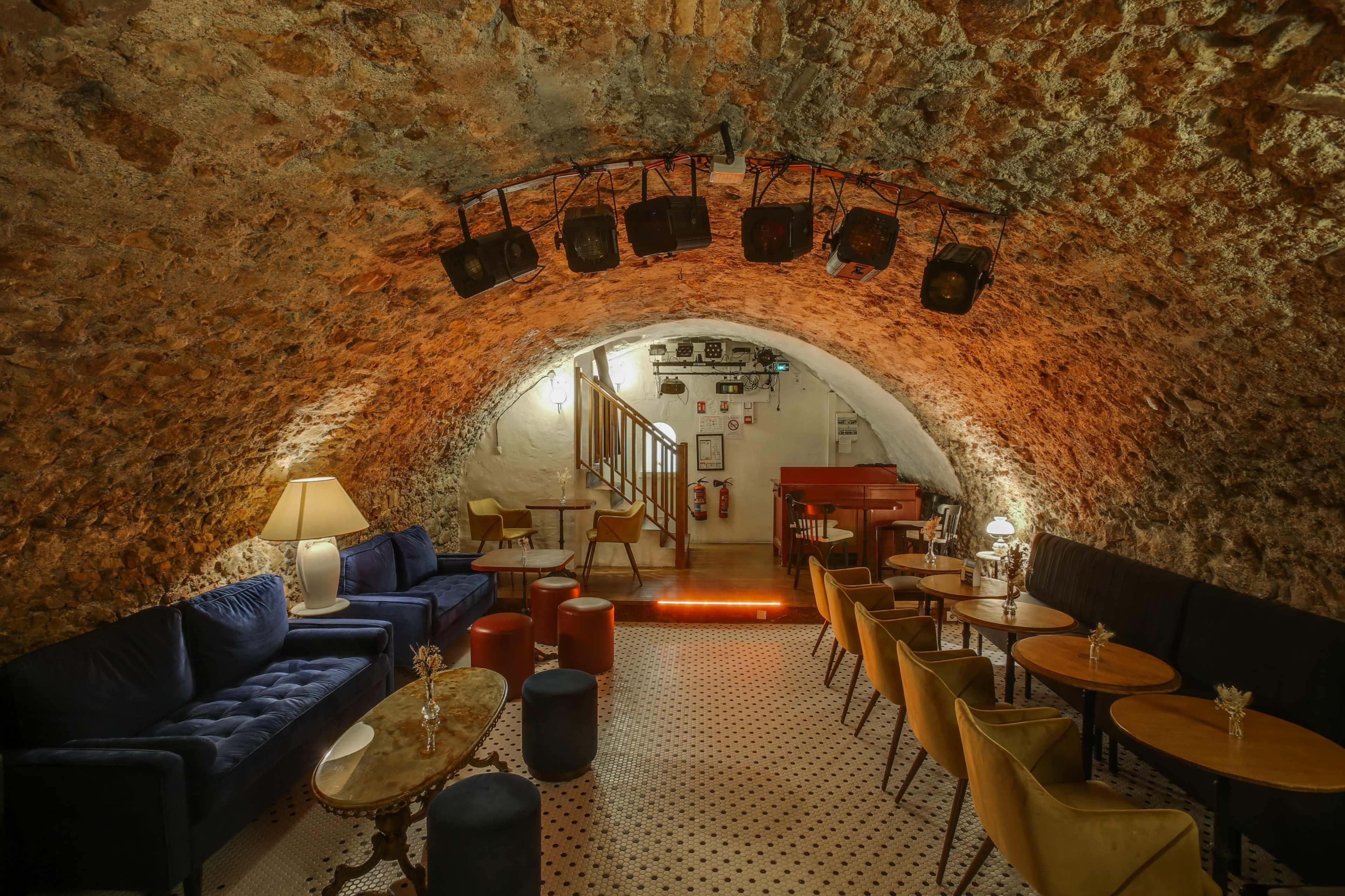 The image shows a cozy underground lounge with stone walls, soft seating, and dim lighting.