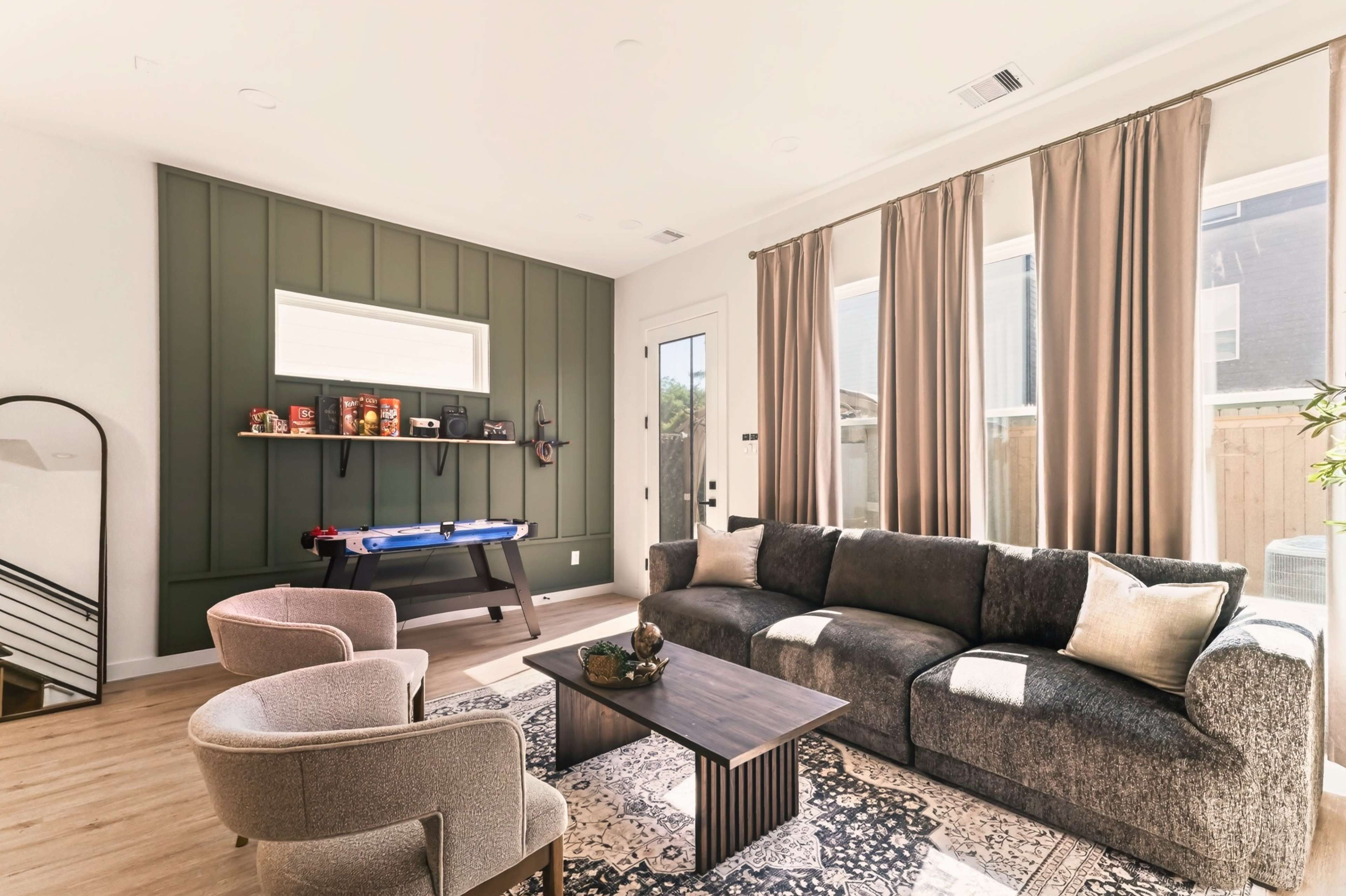 The living room features a gray sofa, two light-colored chairs, a coffee table, and a game table against a green accent wall, with large windows allowing natural light.