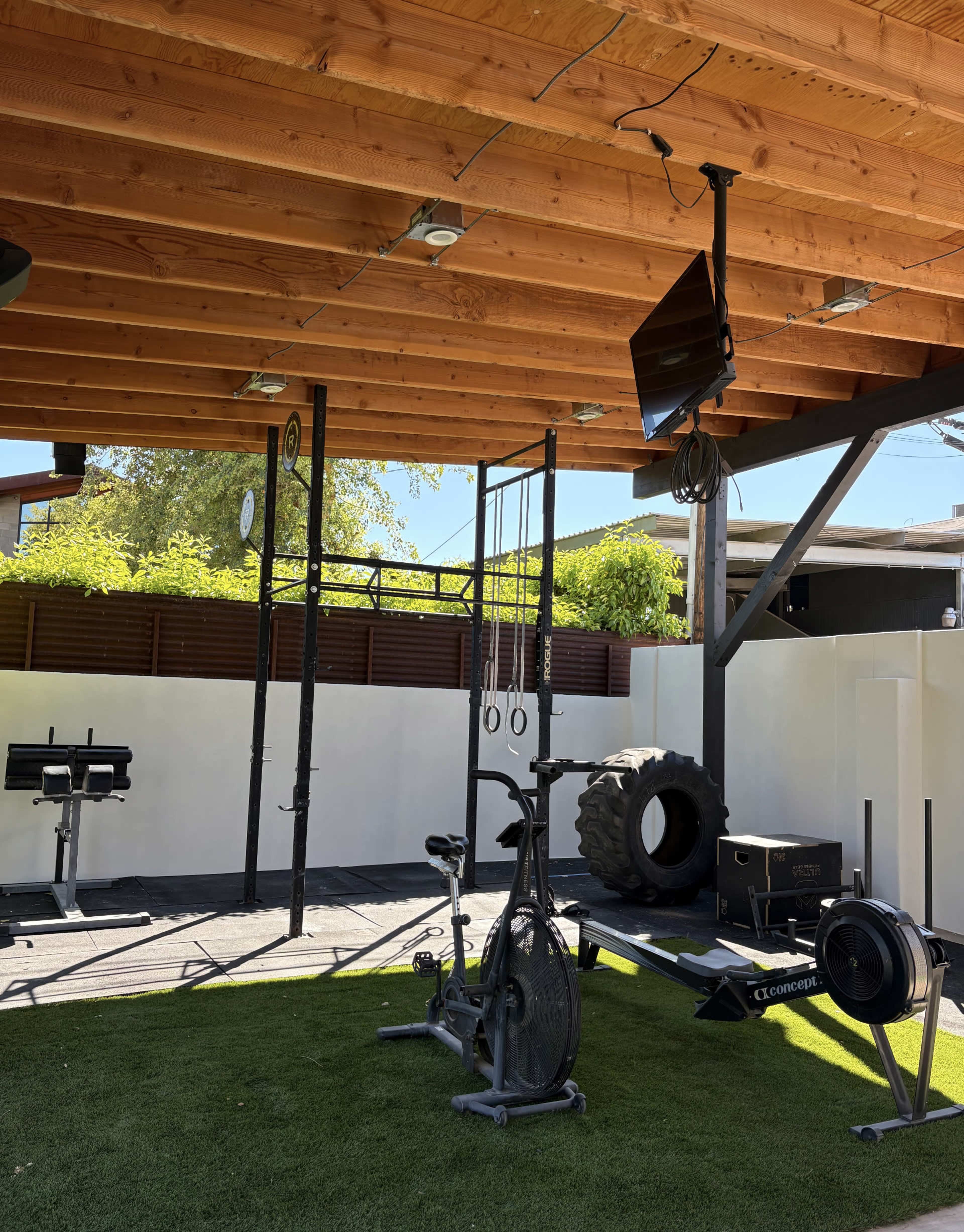 The image shows a spacious outdoor gym with exercise equipment, including a bike, rowing machine, rings, and a large tire, all under a wooden roof.